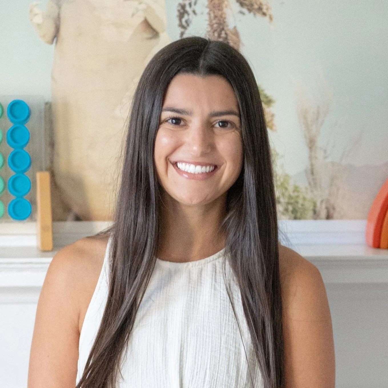 Maddie Abdoney with long dark hair smiling, wearing a sleeveless white top, with a blurred background of decorative objects and a tree painting.