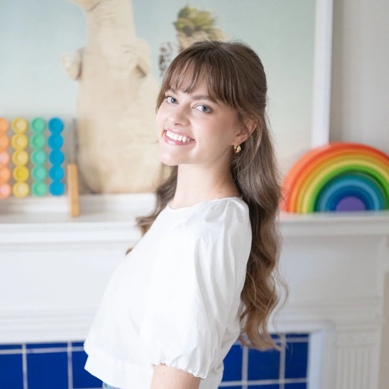 Maya Zwack with long wavy brown hair smiling in a brightly decorated room with a rainbow decoration and colorful abacus in the background.