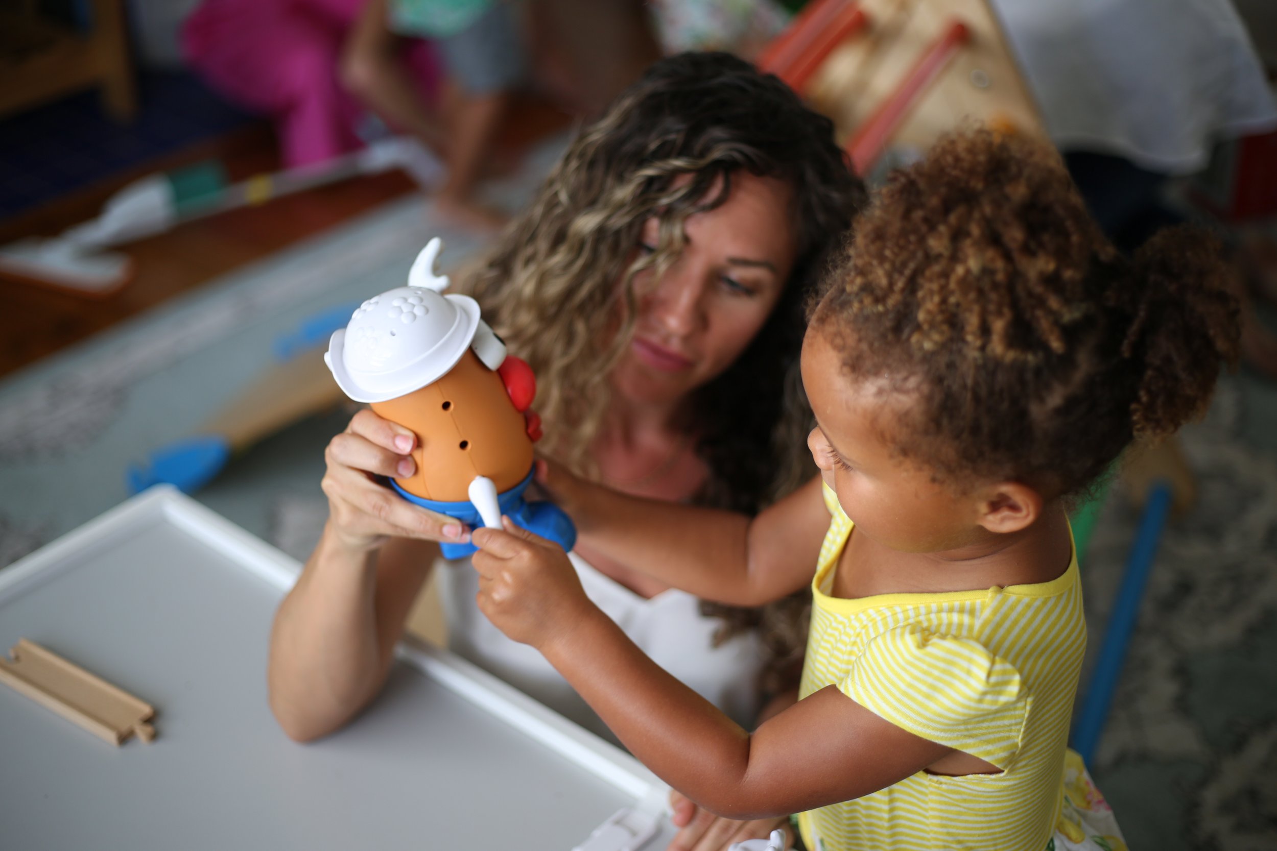 A woman and young girl playing with a toy chef figure in a room filled with toys and books.