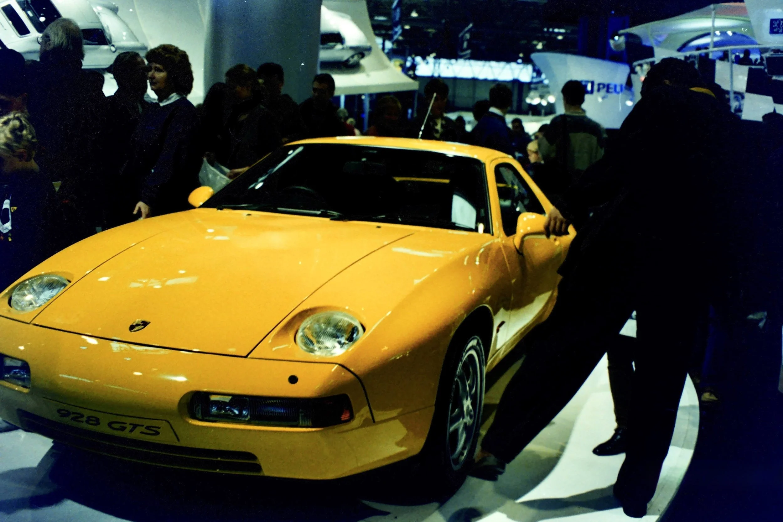 A yellow Porsche 928 GTS sports car displayed at an auto show, surrounded by a crowd of people and other vehicles in the background.
