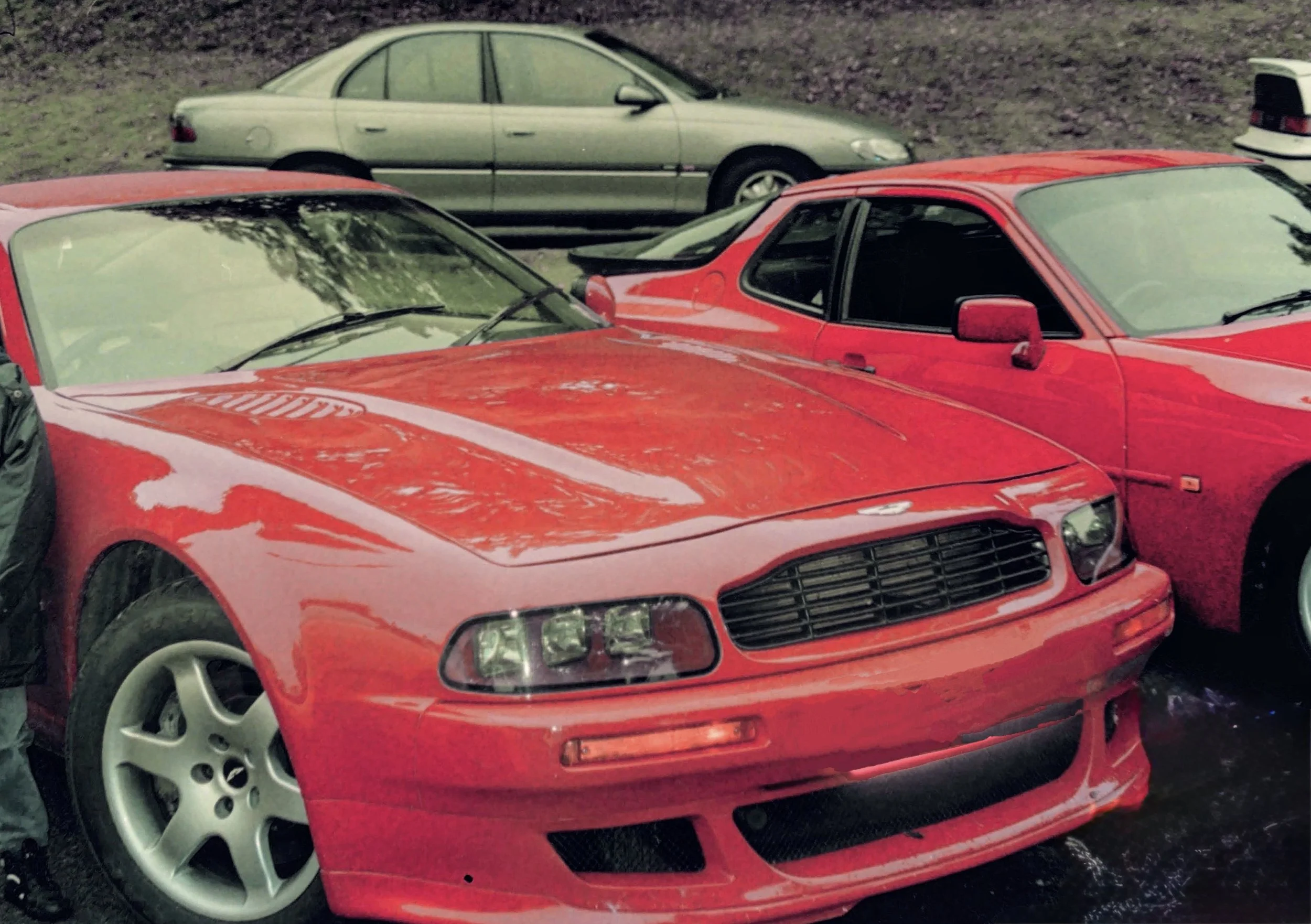 A red sports car parked outdoors, with multiple other cars nearby, against a background of dirt and grass.