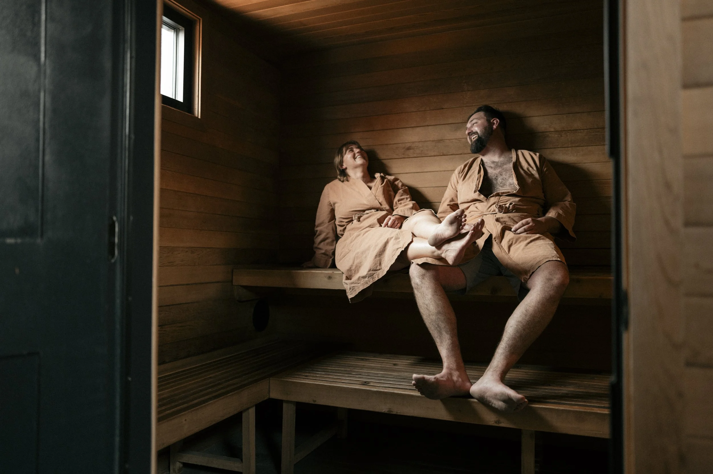 A man and woman sitting inside a wooden sauna, smiling and looking at each other, dressed in tan robes.