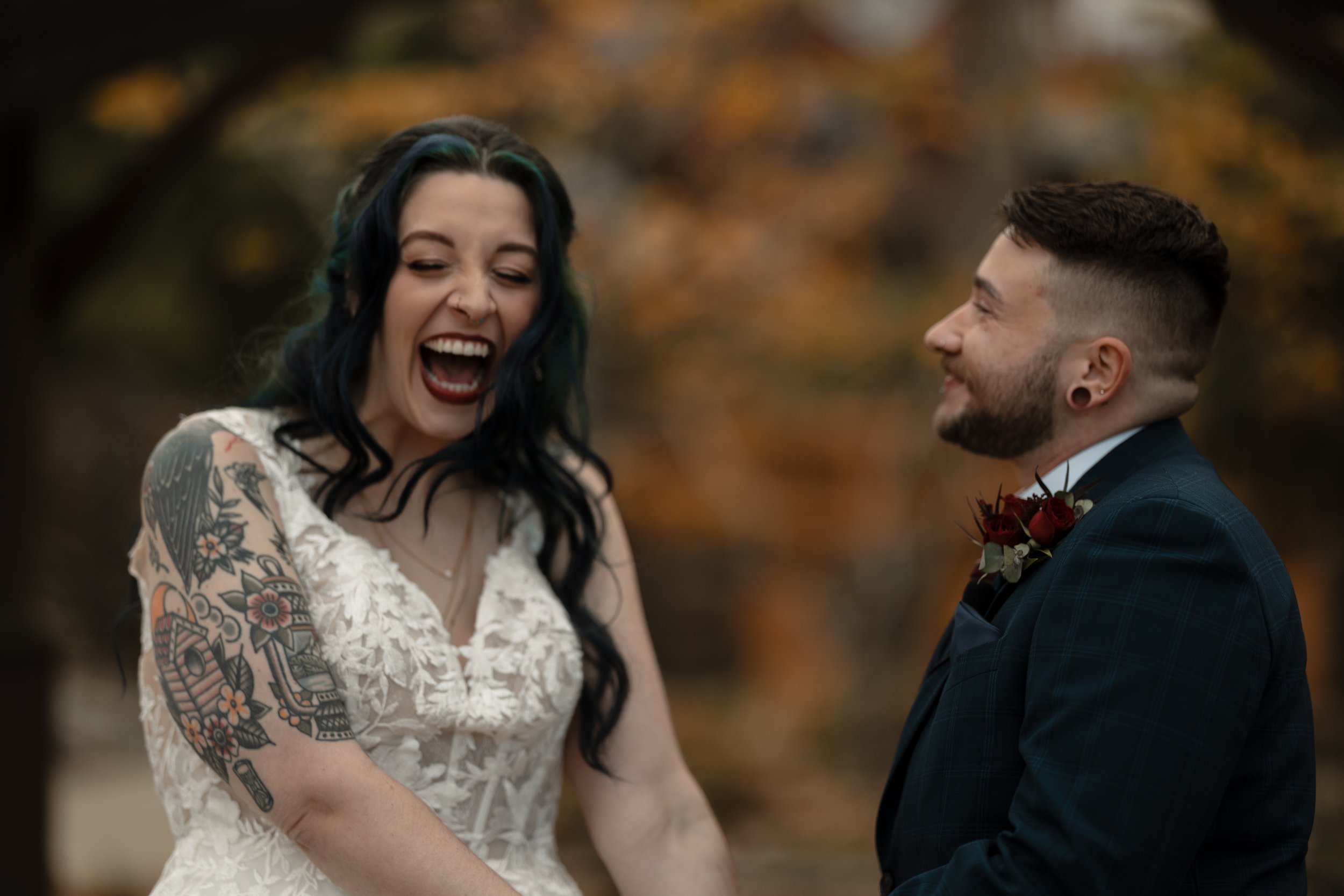 A woman and a man are standing outdoors, smiling and looking at each other, with fall foliage in the background. The woman has black and green hair, tattoos on her arms, and is wearing a white lace dress. The man has short brown hair, earrings, and is dressed in a dark plaid suit with a boutonniere.
