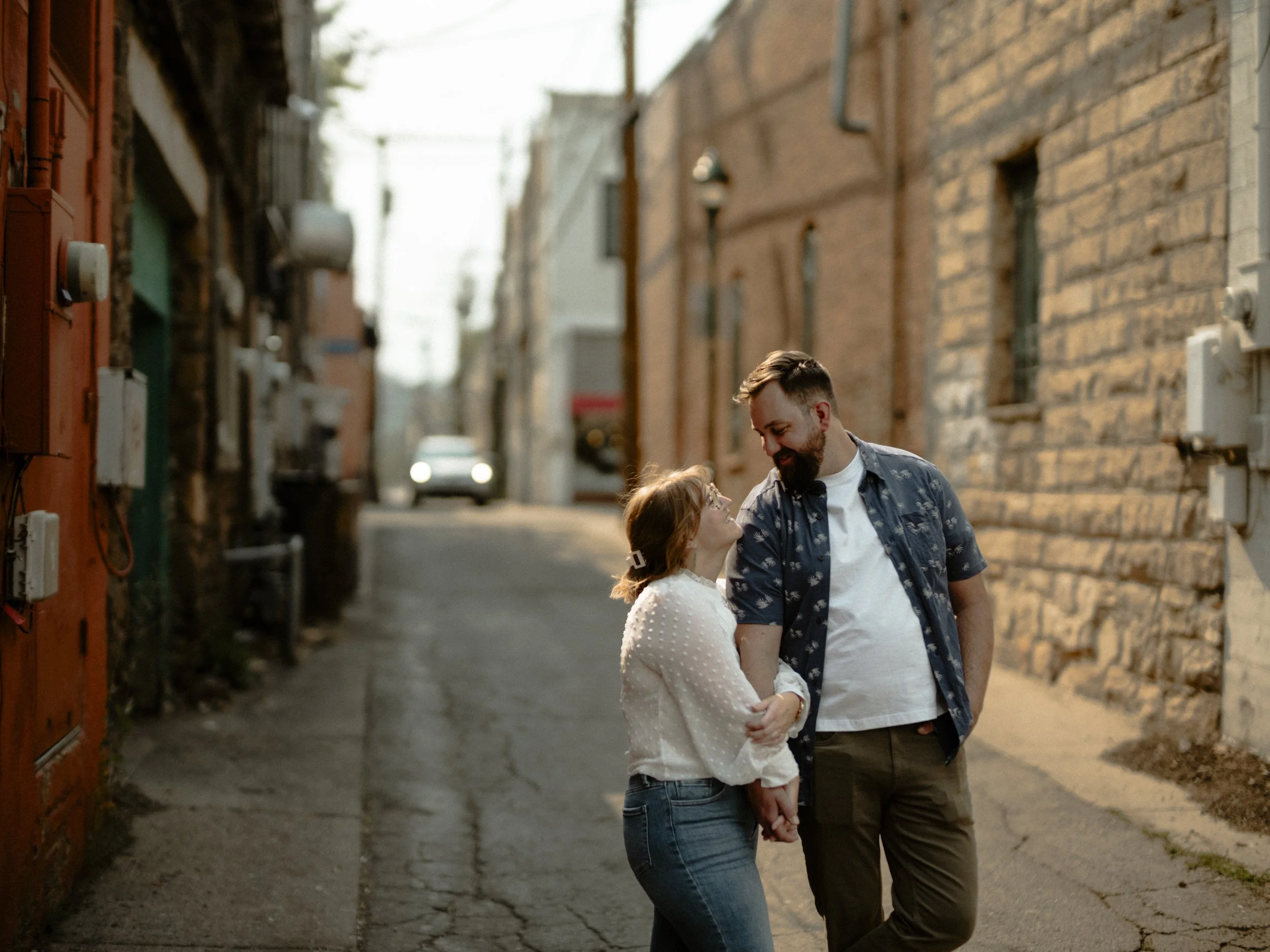A man and woman holding hands and smiling at each other in an alleyway with brick buildings on either side.