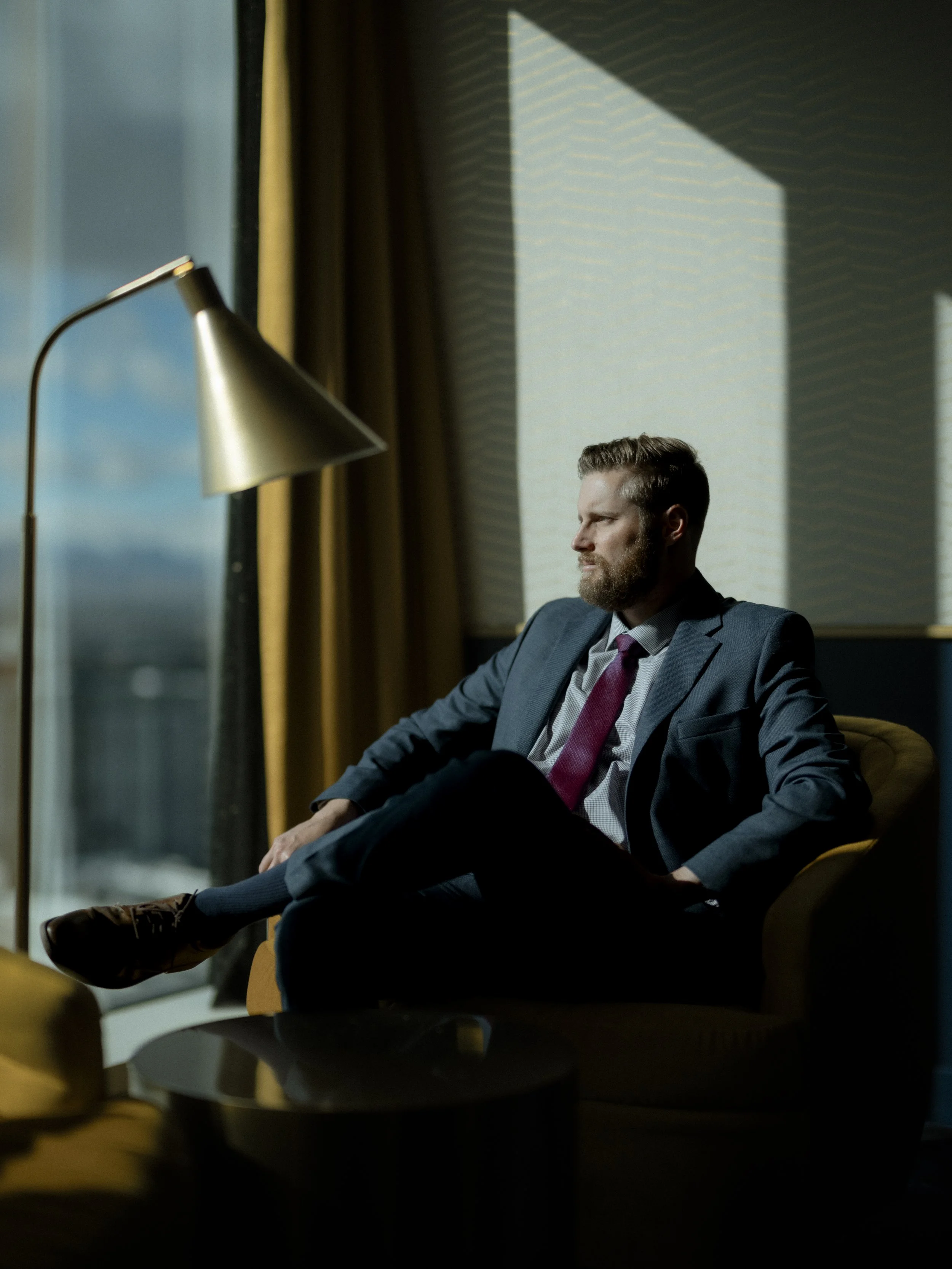 A man in a business suit sitting in a chair near a window, looking thoughtfully outside, with sunlight casting shadows on the wall.