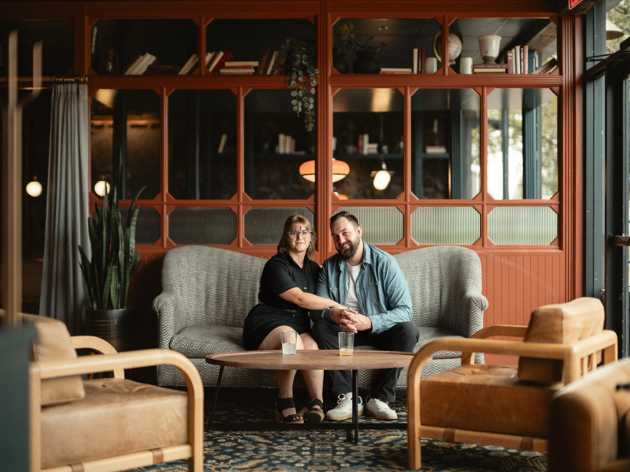 A man and woman sitting close together on a sofa in a cozy, well-lit interior with wood accents, bookshelves, and decorative objects, with a glass of beverage on a nearby table.