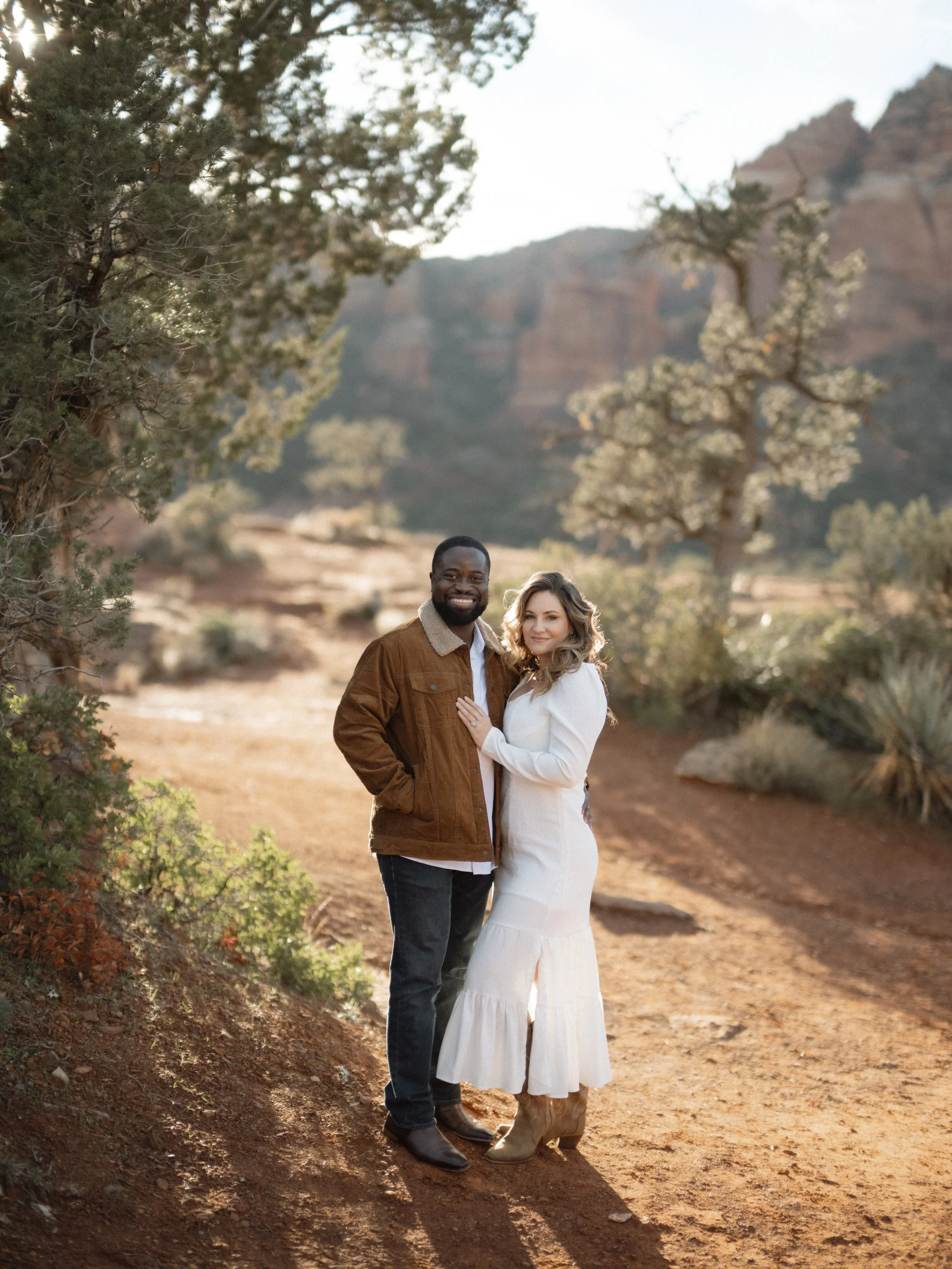 A smiling man and woman stand together on a dirt path in a desert landscape with trees and red rock mesas in the background, during sunset.