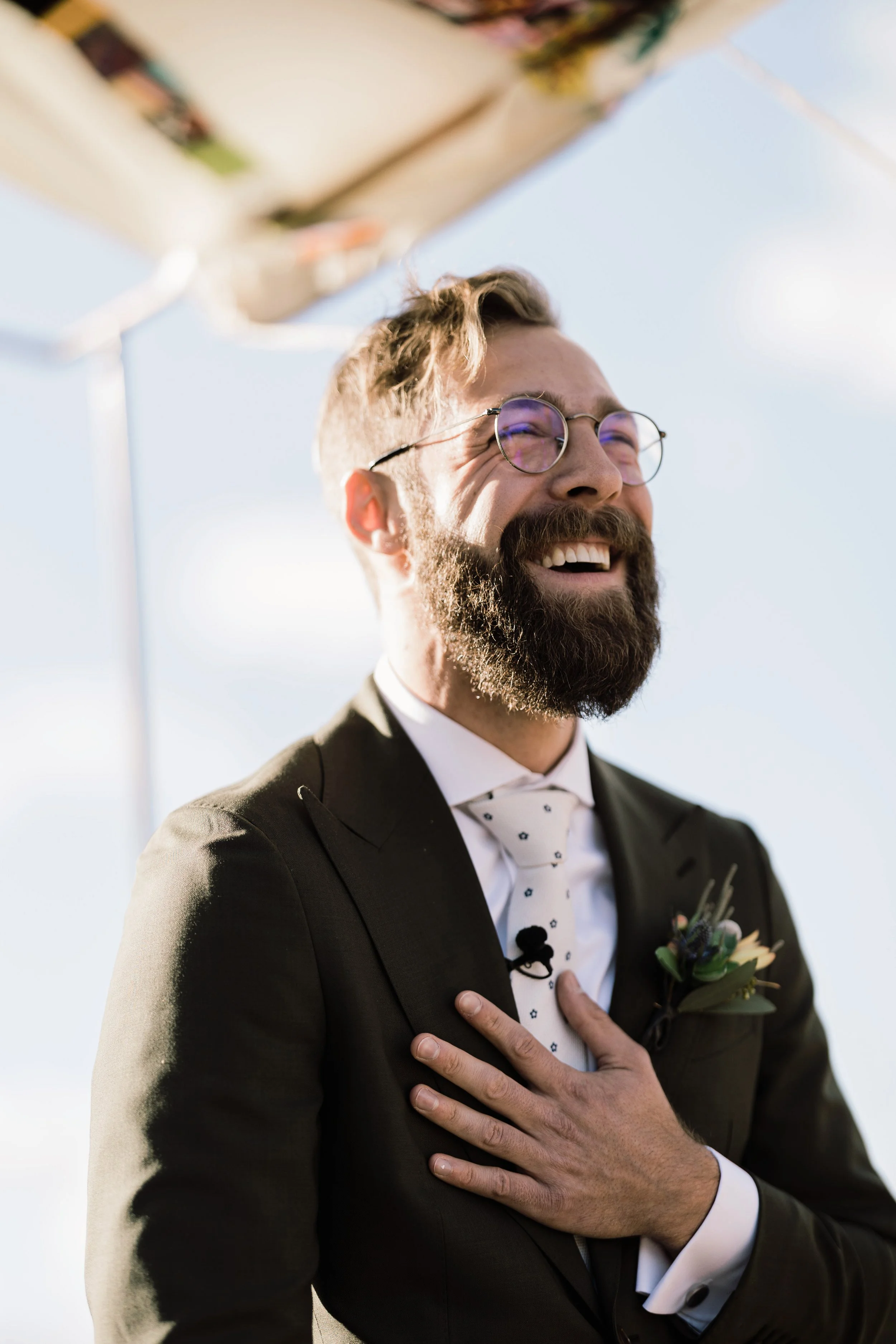 A man in a suit with a boutonniere, wearing glasses, smiling with his eyes closed, with his left hand over his heart, under a partly cloudy sky.