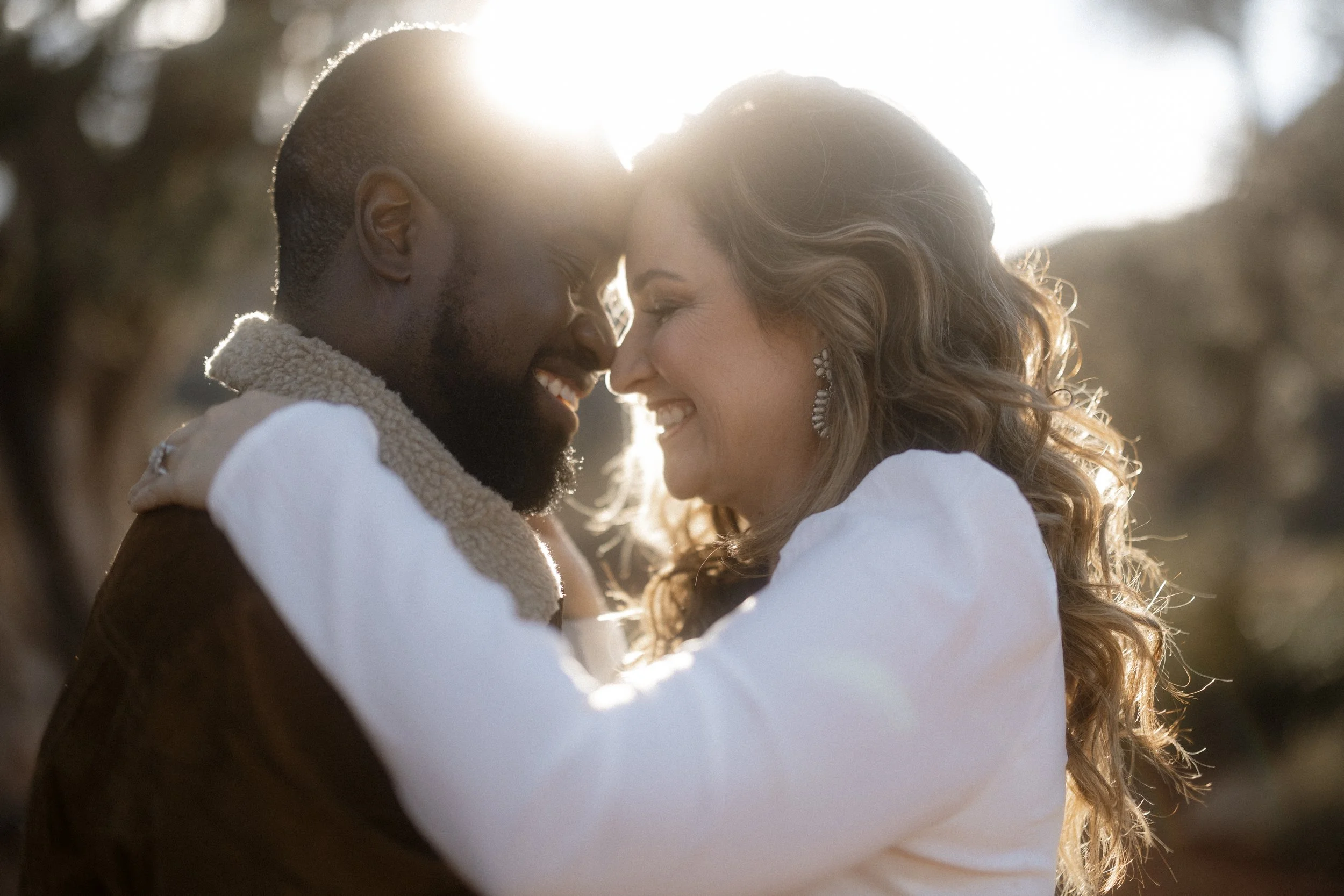 A happy couple embracing outdoors during sunset, touching foreheads and smiling, with sunlight behind them.
