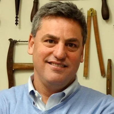 A man with short graying hair smiling at the camera, wearing a light blue sweater over a collared shirt, standing in front of a wall with various woodworking tools hanging on it.