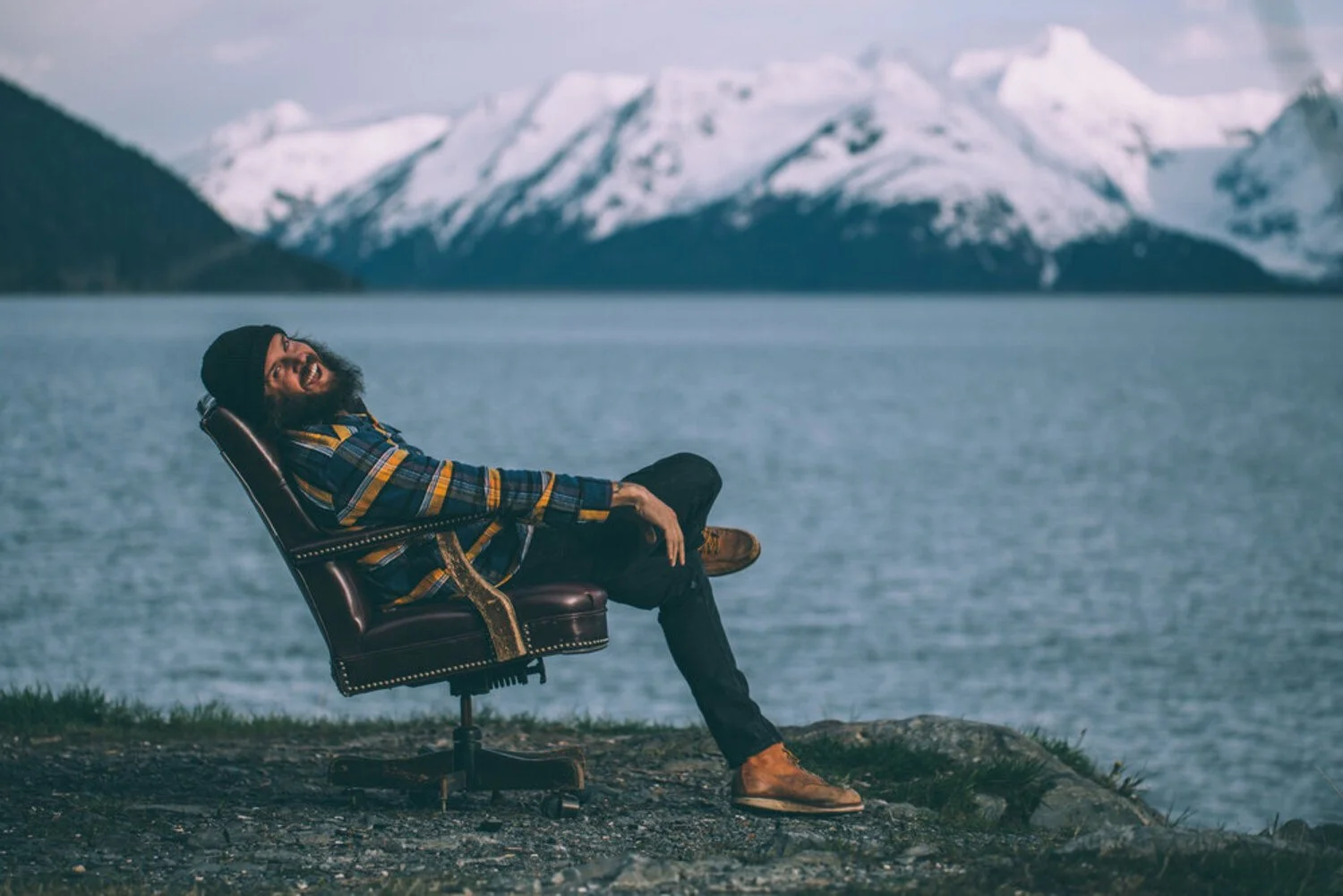 A man with glasses and a beard sitting relaxed in a chair outdoors near water, with snow-capped mountains in the background.