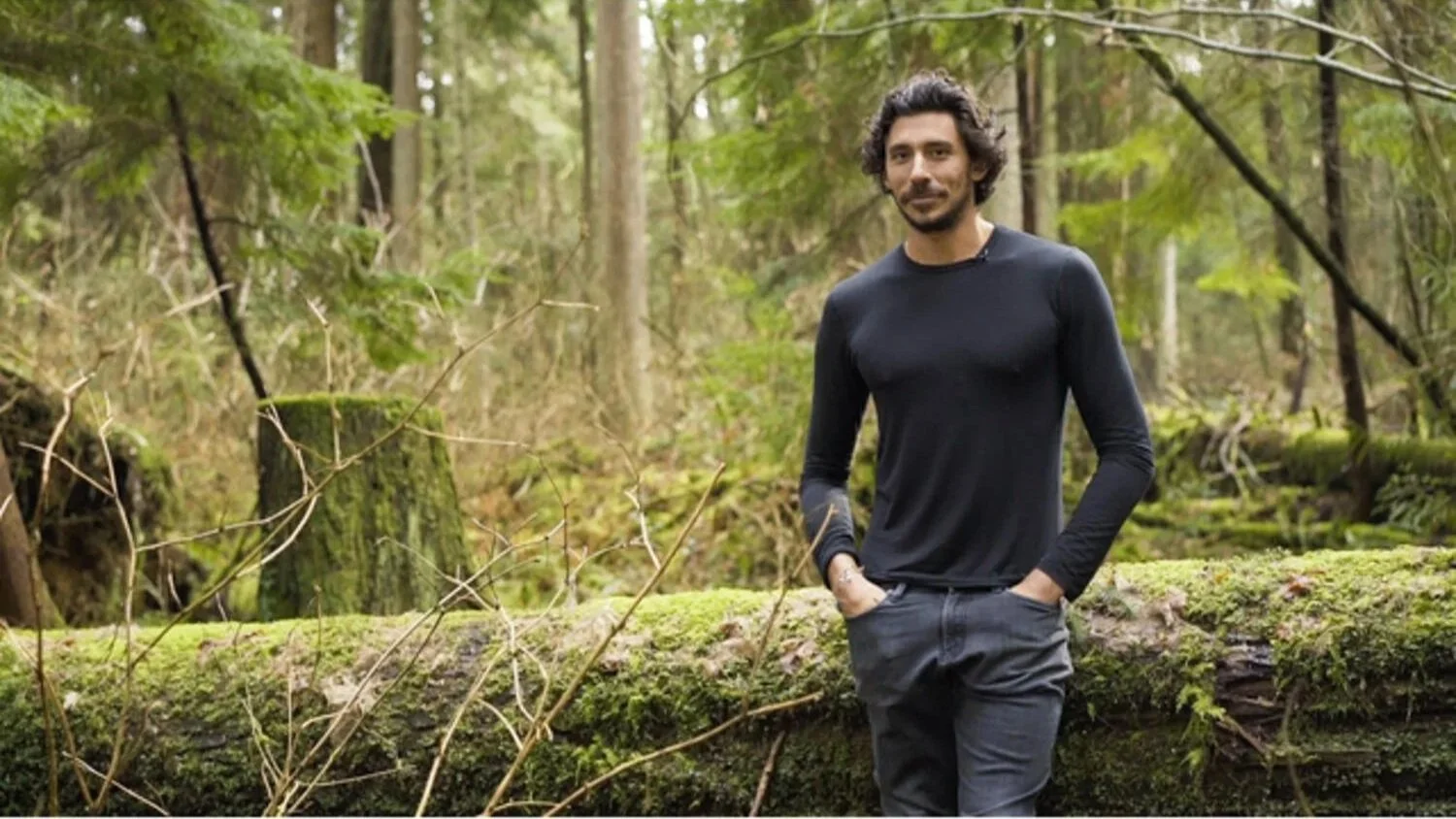 A man with curly hair and a beard standing in a forest, wearing a black long-sleeve shirt and gray pants, with hands in pockets, surrounded by trees and greenery.