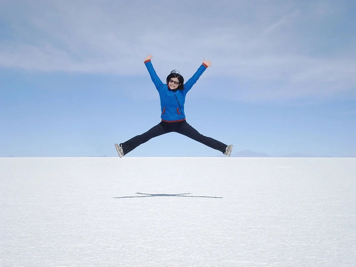 A woman jumping in the air with arms and legs spread wide on a snowy landscape under a partly cloudy sky.