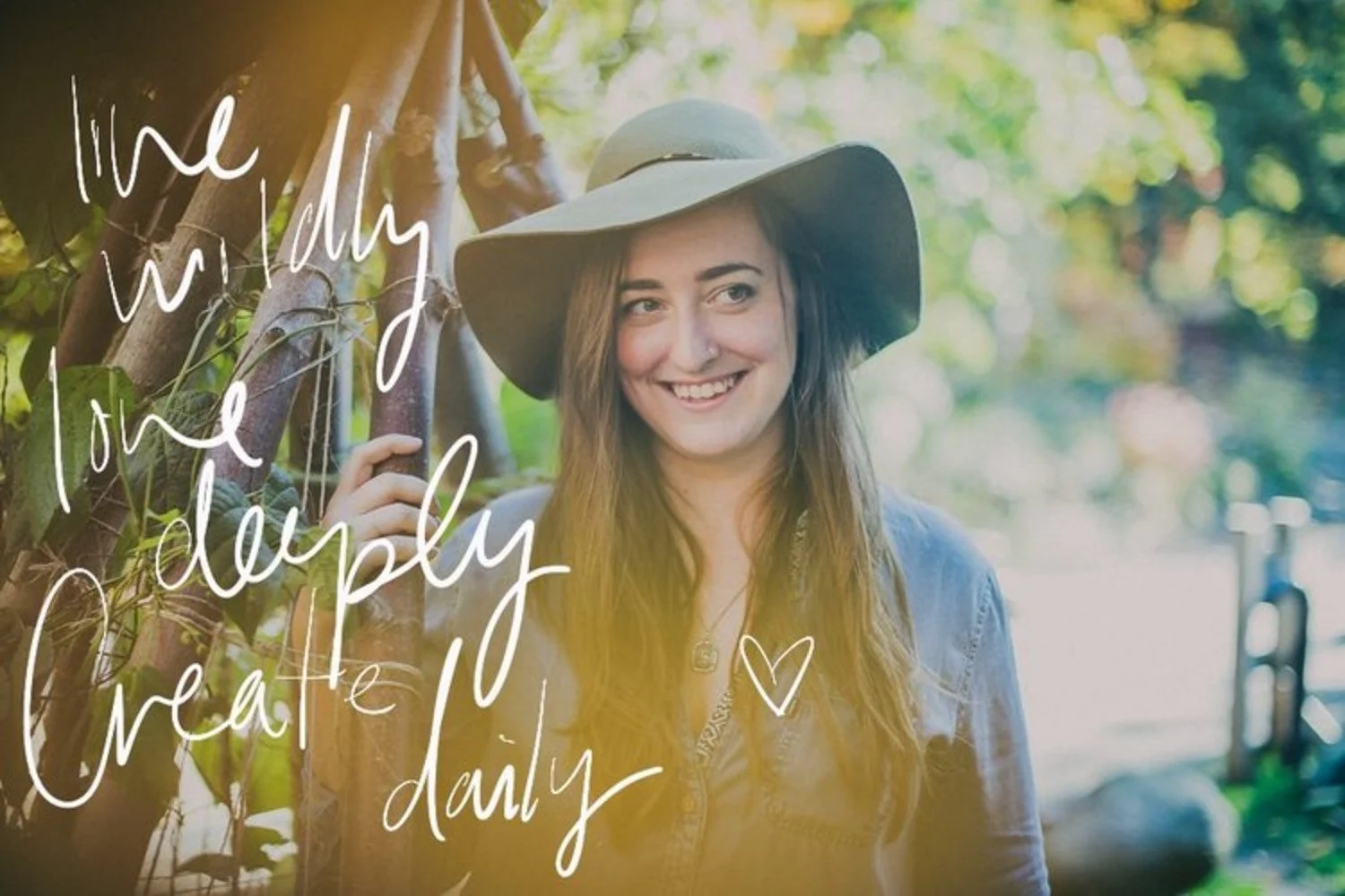 A woman with long brown hair wearing a wide-brimmed hat, smiling outdoors in a sunny park with trees and a walkway in the background.