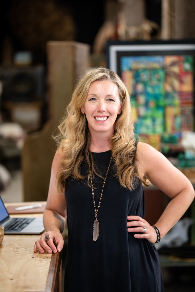 A smiling woman with long, wavy blonde hair wearing a black sleeveless dress and a long feather necklace, standing in a cozy indoor space with artworks and a laptop on a wooden table.