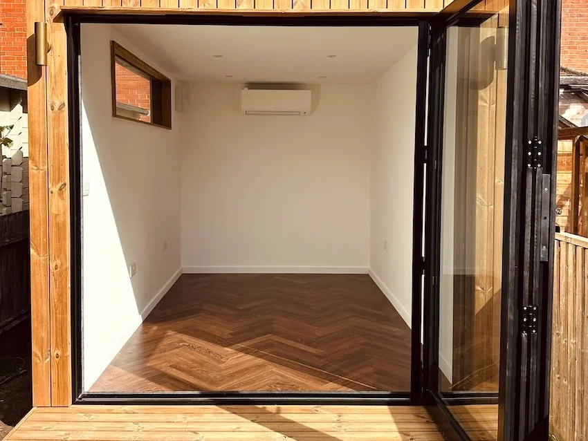 Interior of a bespoke garden office with chevron flooring, air conditioning and open bi-fold doors.