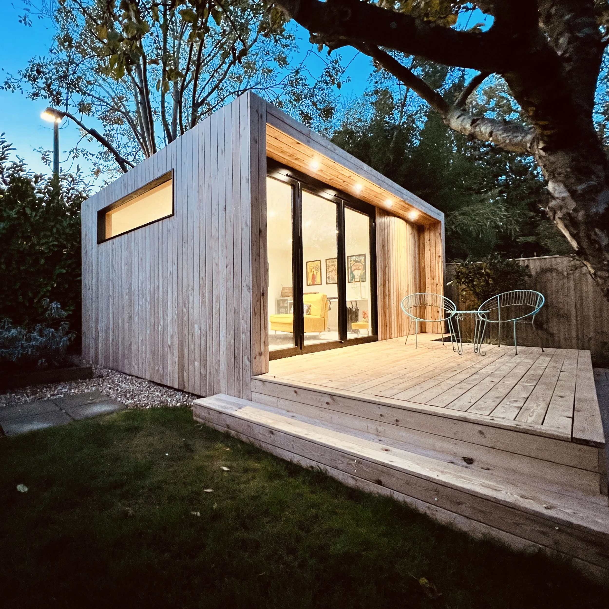 Angled exterior view of the LUNA 5m x 4m garden room with decking, shown at dusk within a landscaped garden