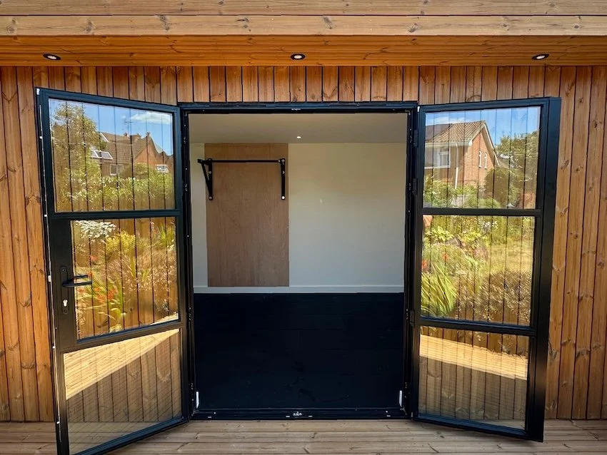 Bespoke garden gym with open black steel-framed glazed French doors and a covered timber deck.