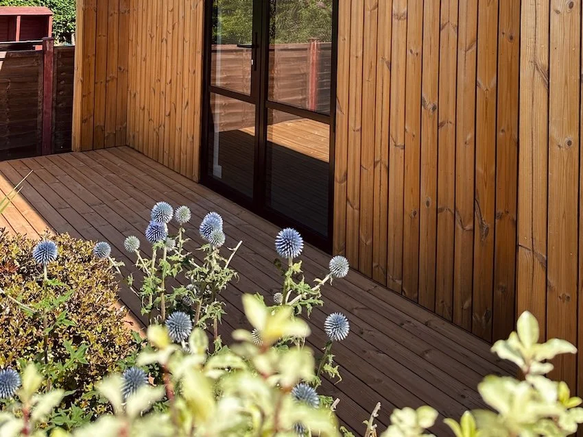 Timber-clad bespoke garden gym with black glazed doors, a covered deck and established garden planting.