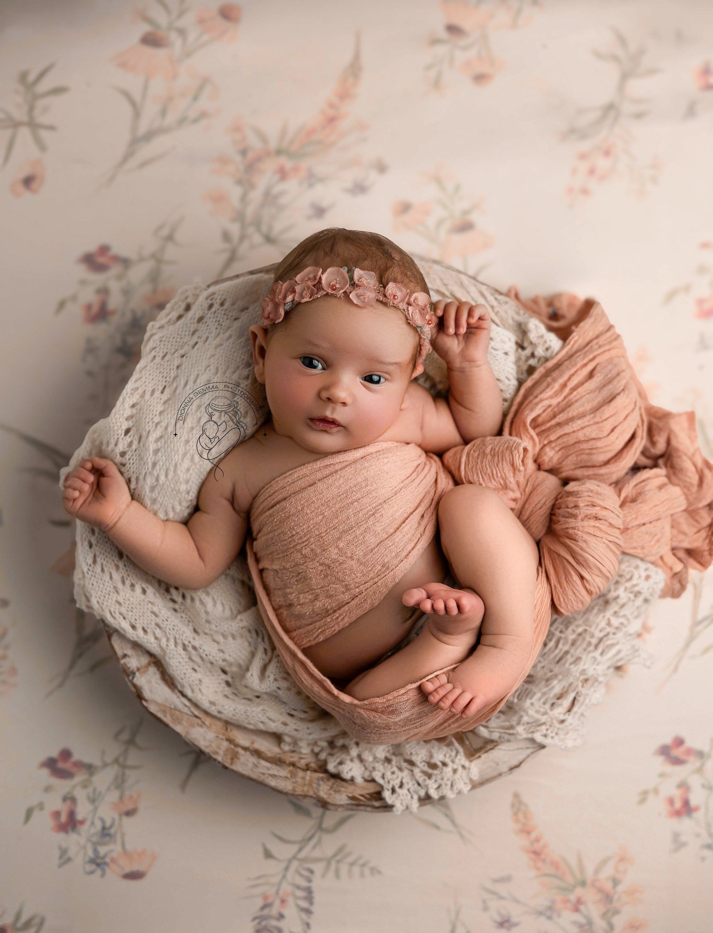 A baby girl with blue eyes, wearing a pink headband with fabric flowers, lying on a crochet blanket in a woven basket, surrounded by pink and peach-colored fabric, with a floral background.