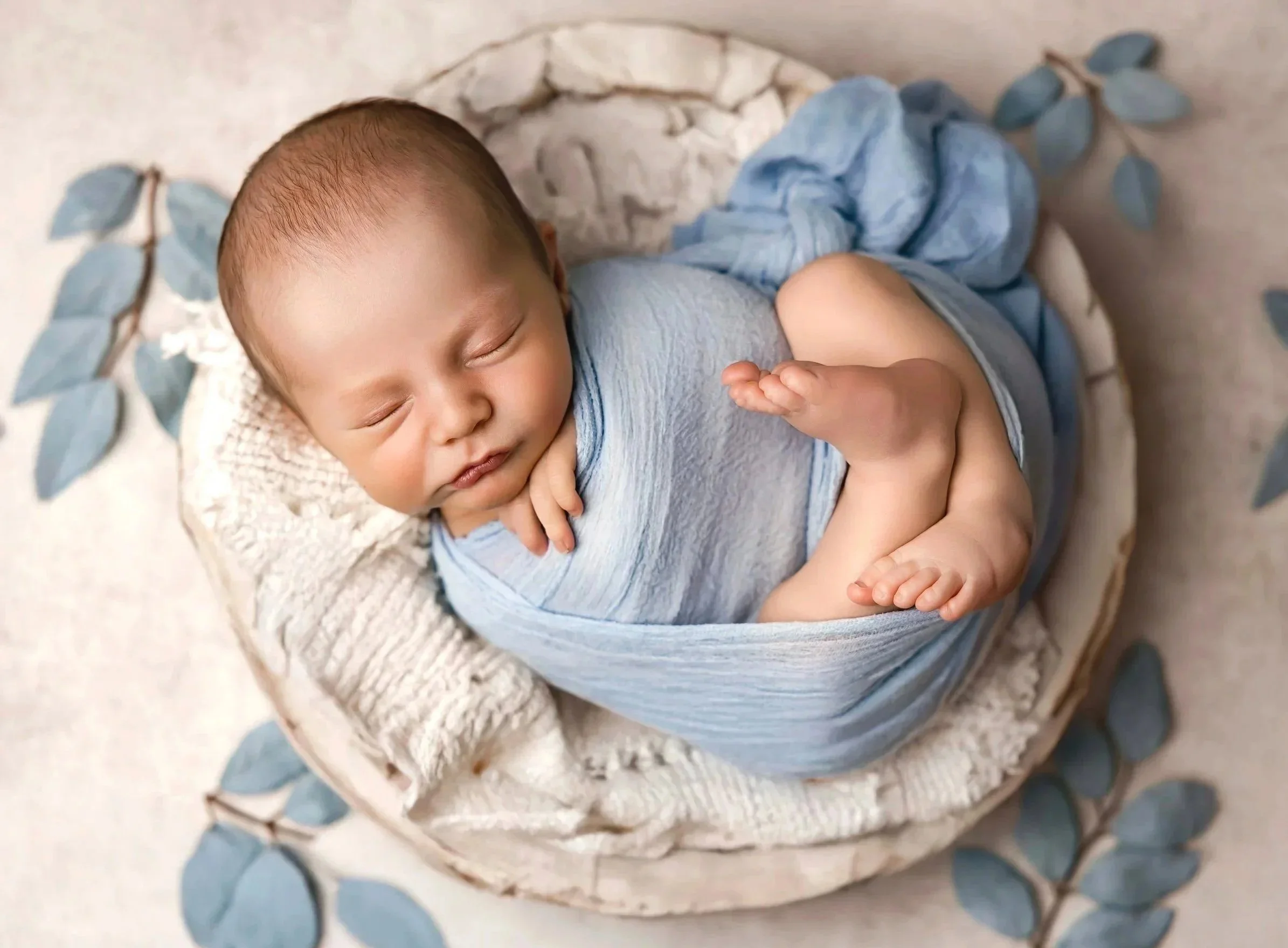 A sleeping baby dressed in a light blue outfit, lying on a small bed with a beige knitted blanket. The bed has a textured off-white pillow and is decorated with a branch of blue leaves around it.