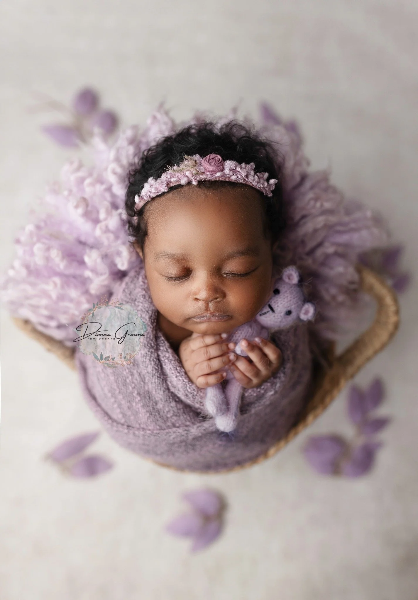 A baby with curly hair wearing a pink floral headband and a purple knitted sweater, sleeping with hands clasped together, surrounded by purple petals.