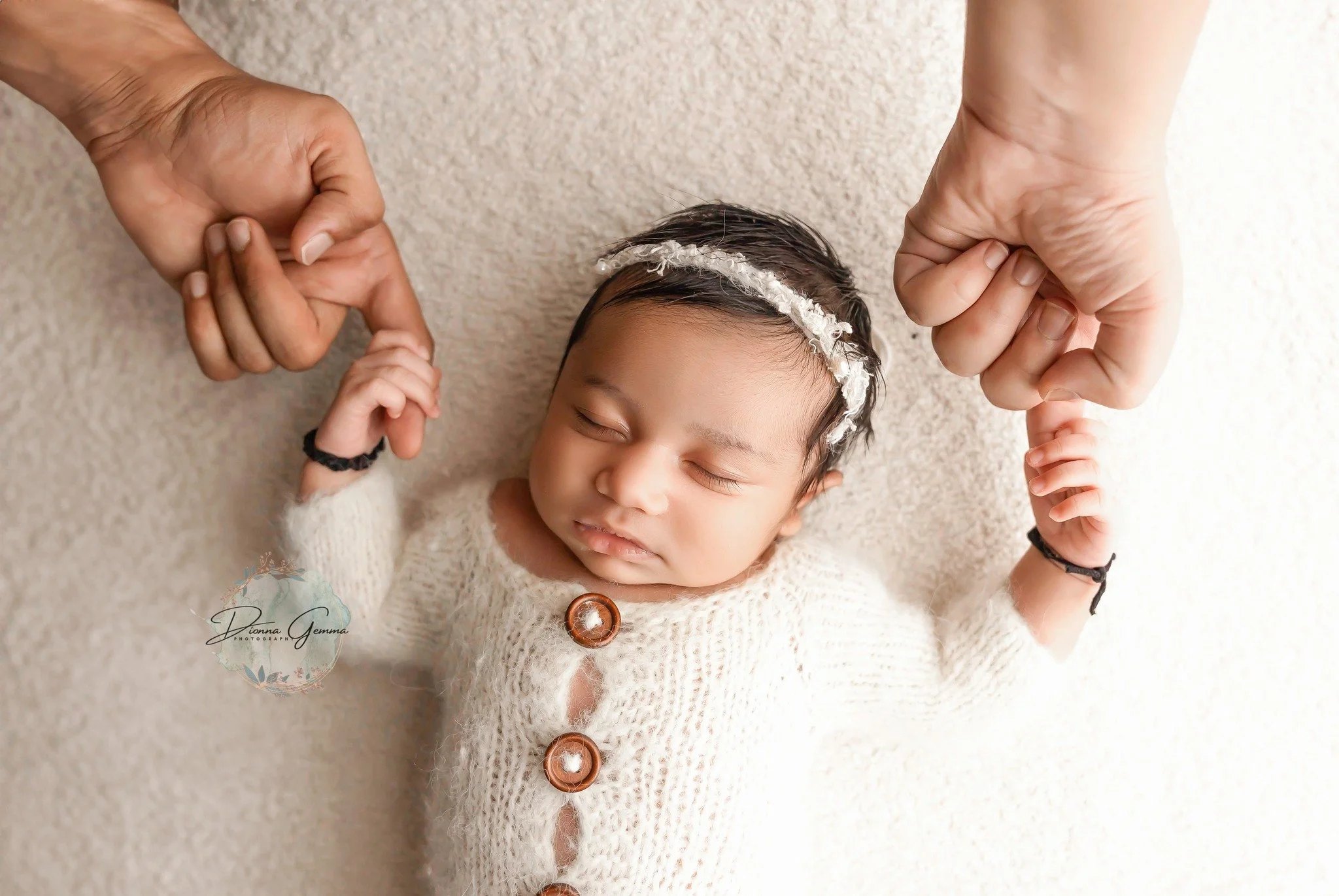 A baby girl with dark hair, wearing a white sweater with brown buttons and a headband, lies on a light-colored carpet. Two hands hold her tiny hands, one on each side of her. The baby appears to be sleeping peacefully.