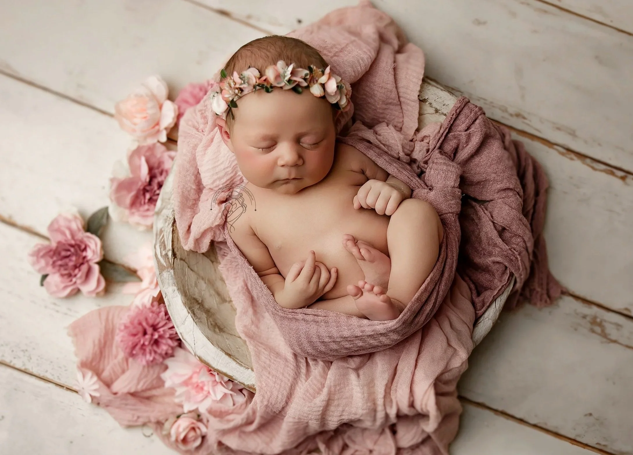 A sleeping baby girl with a flower crown on her head, wrapped in pink fabric, lying on a pink blanket inside a rustic wooden bowl surrounded by pink flowers.