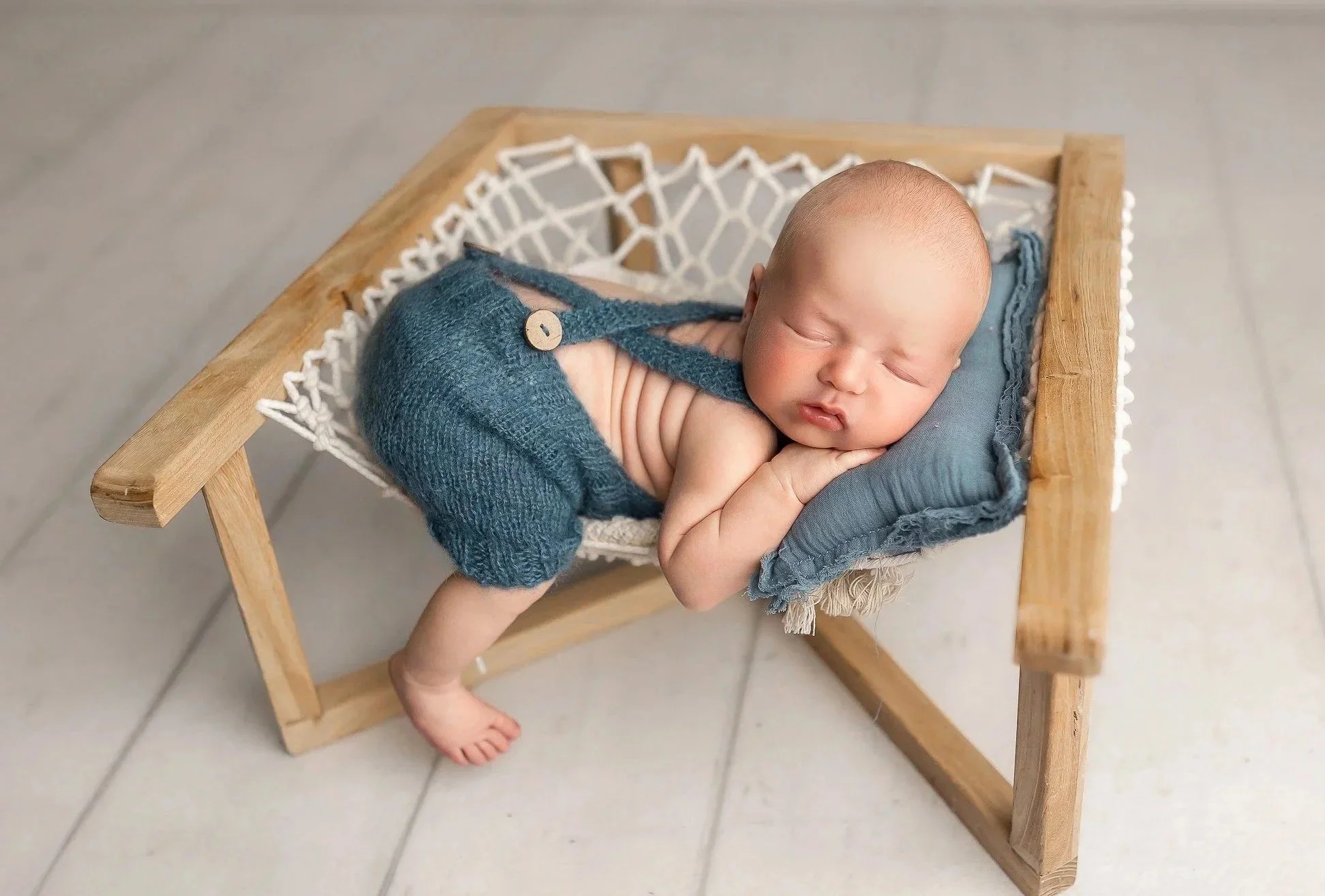 A sleeping baby lying on his side in a small wooden bed, with a blue blanket and a blue knitted outfit with a button on it, on a tiled floor.