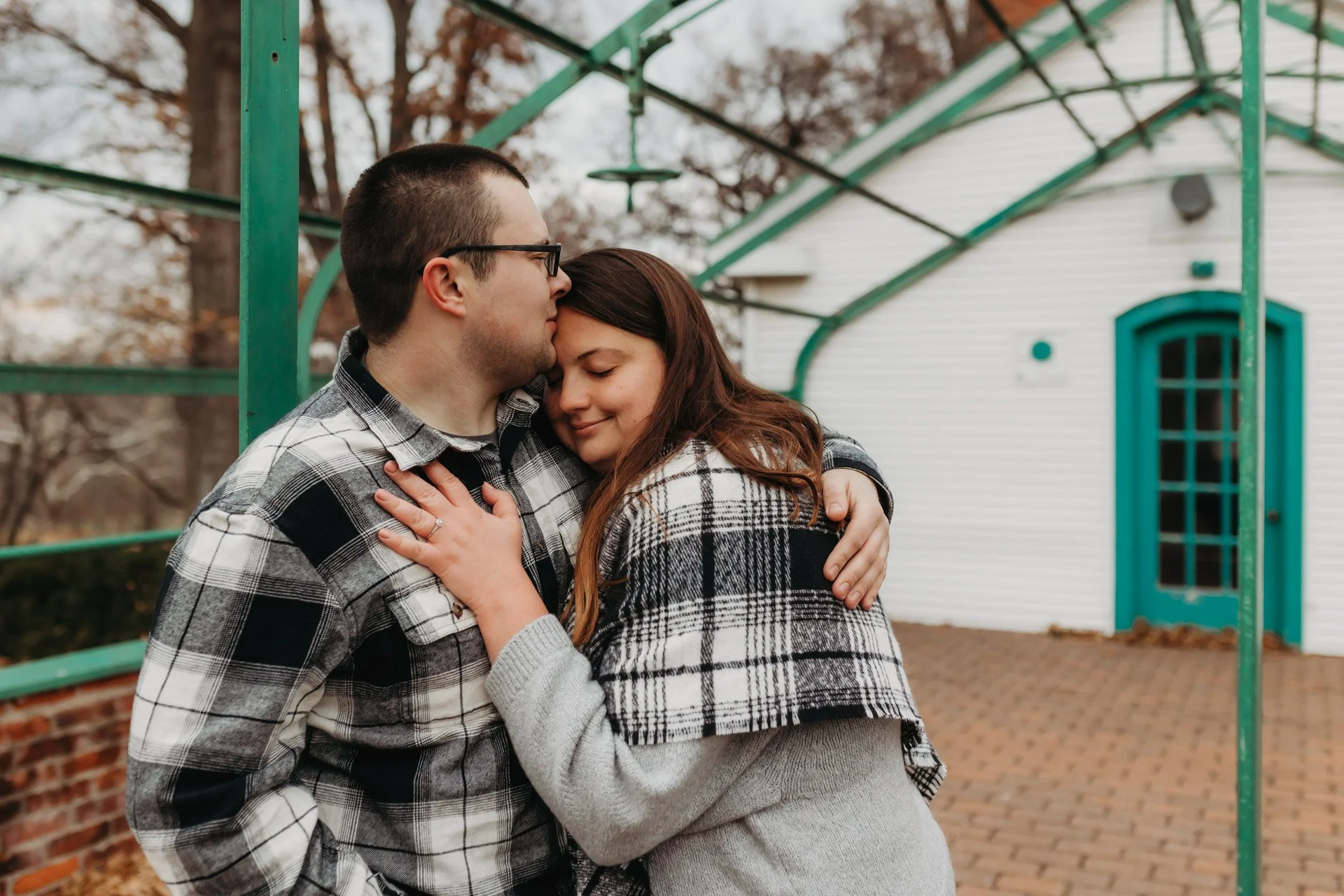 A couple embracing outdoors in front of a white building with teal accents during fall, with trees in the background at their engagement photography session at Queeny Park in STL.