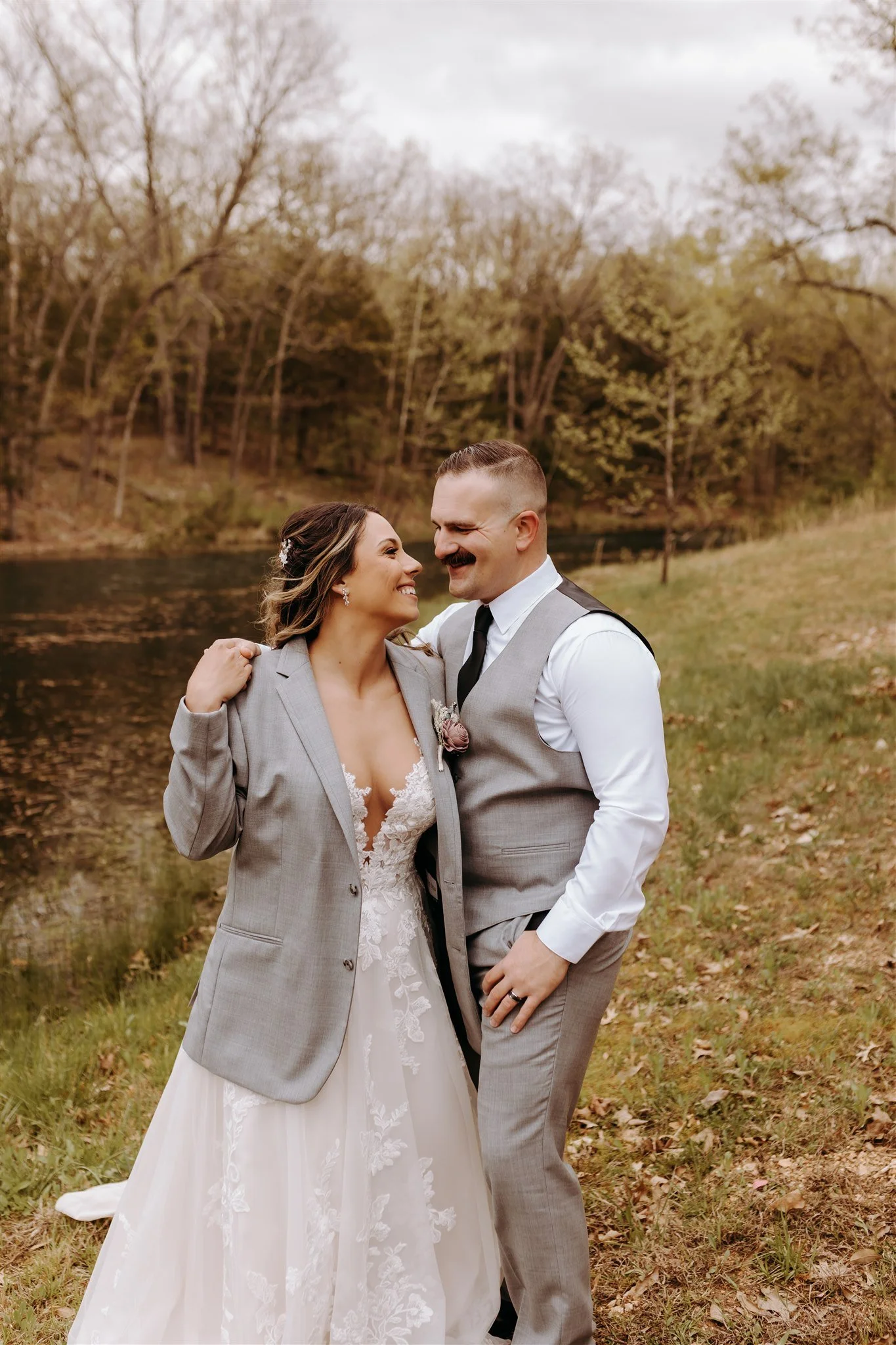 A newlywed couple in wedding attire stands close together outdoors near a river at Rolla, MO rustic wedding venue, The Village. Photos by Columbia, MO Photographer Liv Strange Photography.