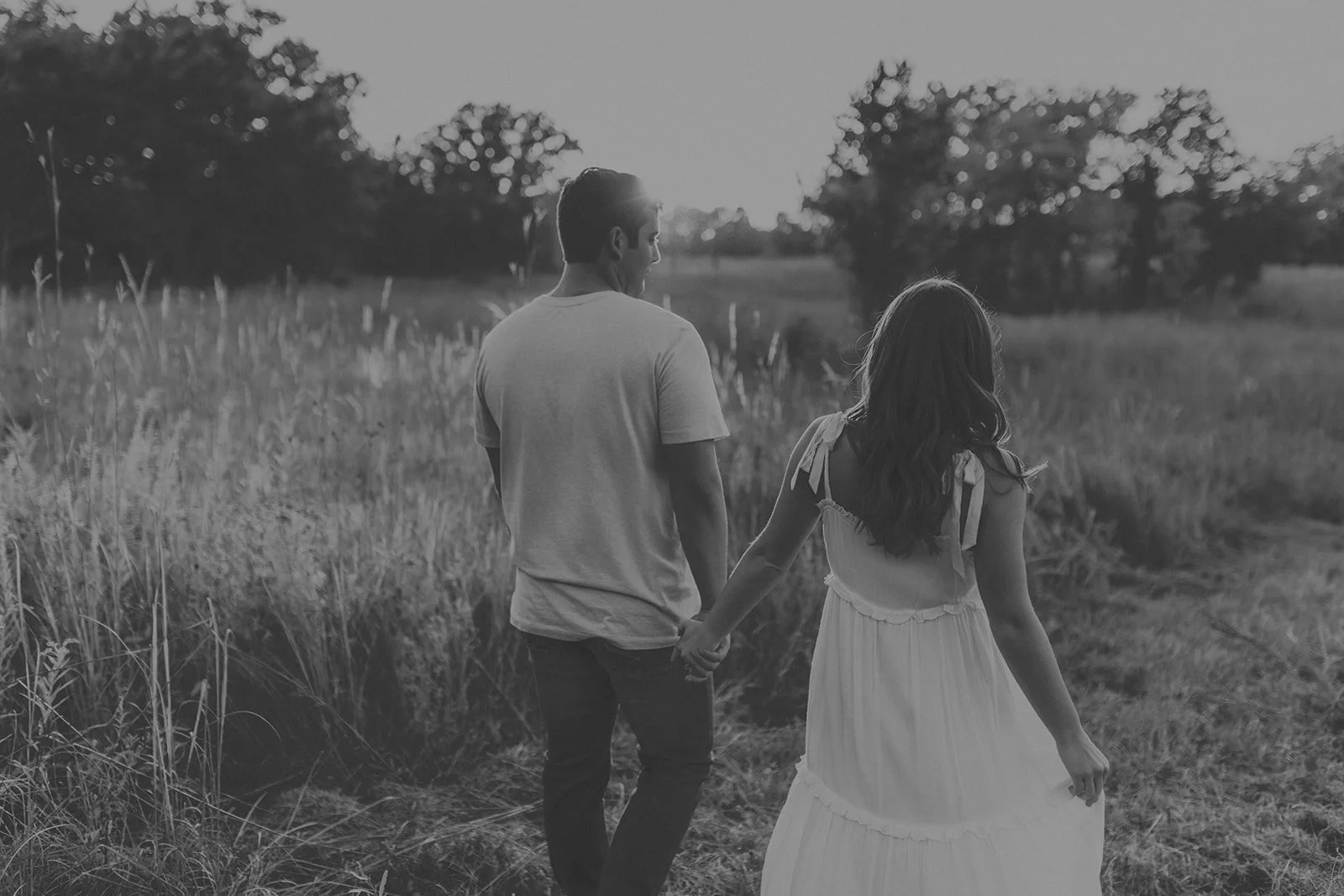 A black-and-white photo of a man and woman walking hand-in-hand in a field during sunset or sunrise during their engagement photography session at Rock Bridge State Park in Columbia, MO.