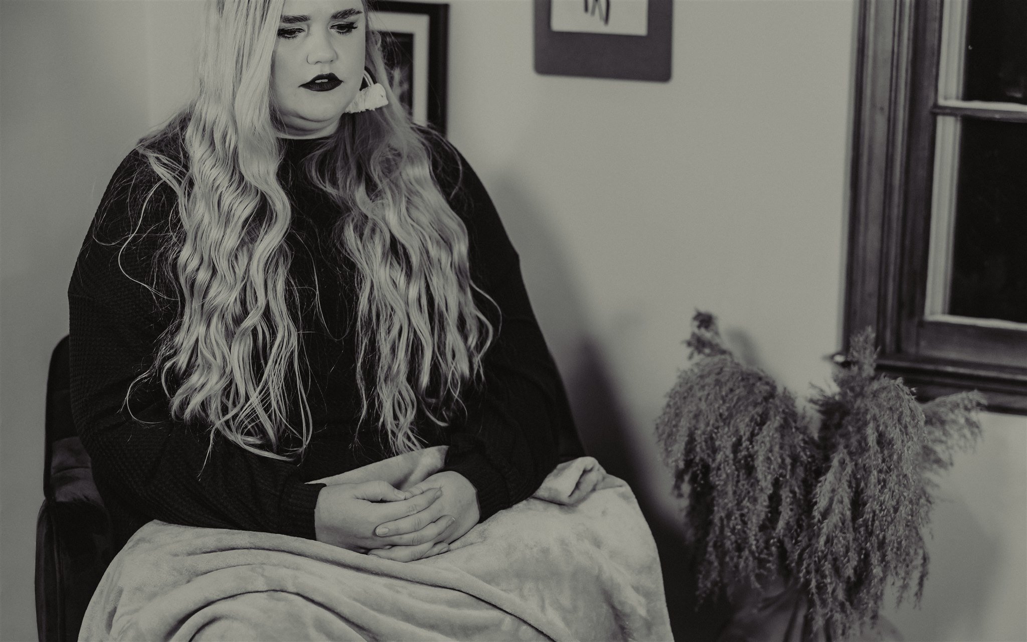 A woman with long wavy hair and dark lipstick sitting on a chair, with her hands clasped in her lap, beside a textured potted plant during a trauma informed photography session in Columbia, Missouri.