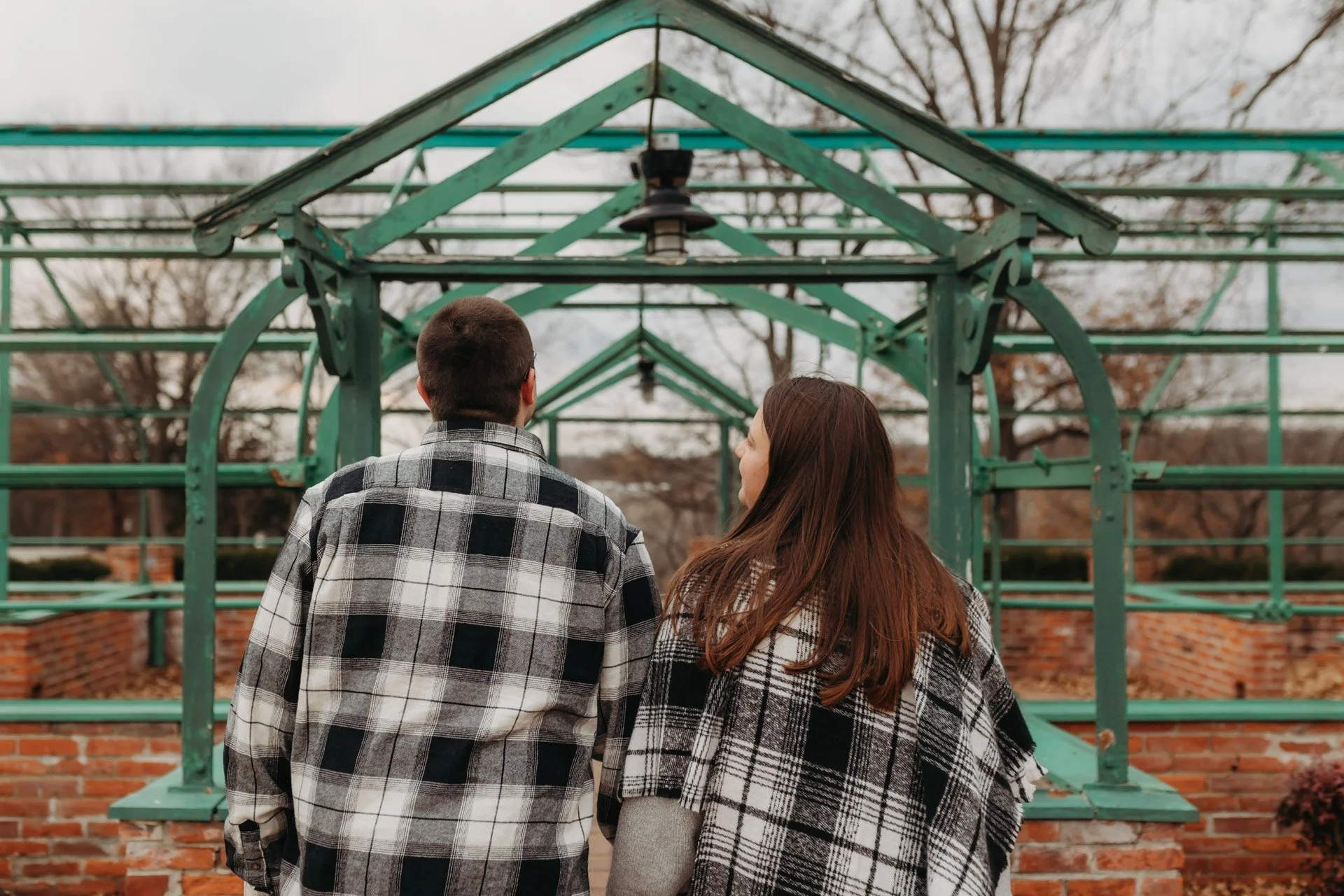 A young man and woman with their backs to the camera, dressed in plaid shirts, walking through an outdoor metal and brick tunnel structure in an urban park at their engagement photography session at Queeny Park in STL.
