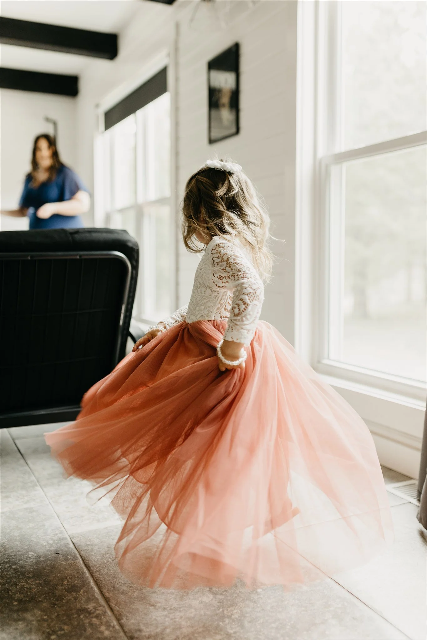 A young flower girl in a white lace top and a peach tutu dress twirling indoors near large windows at Rolla, Missouri rustic wedding venue, The Village. Photos by Columbia, MO Photographer Liv Strange Photography.