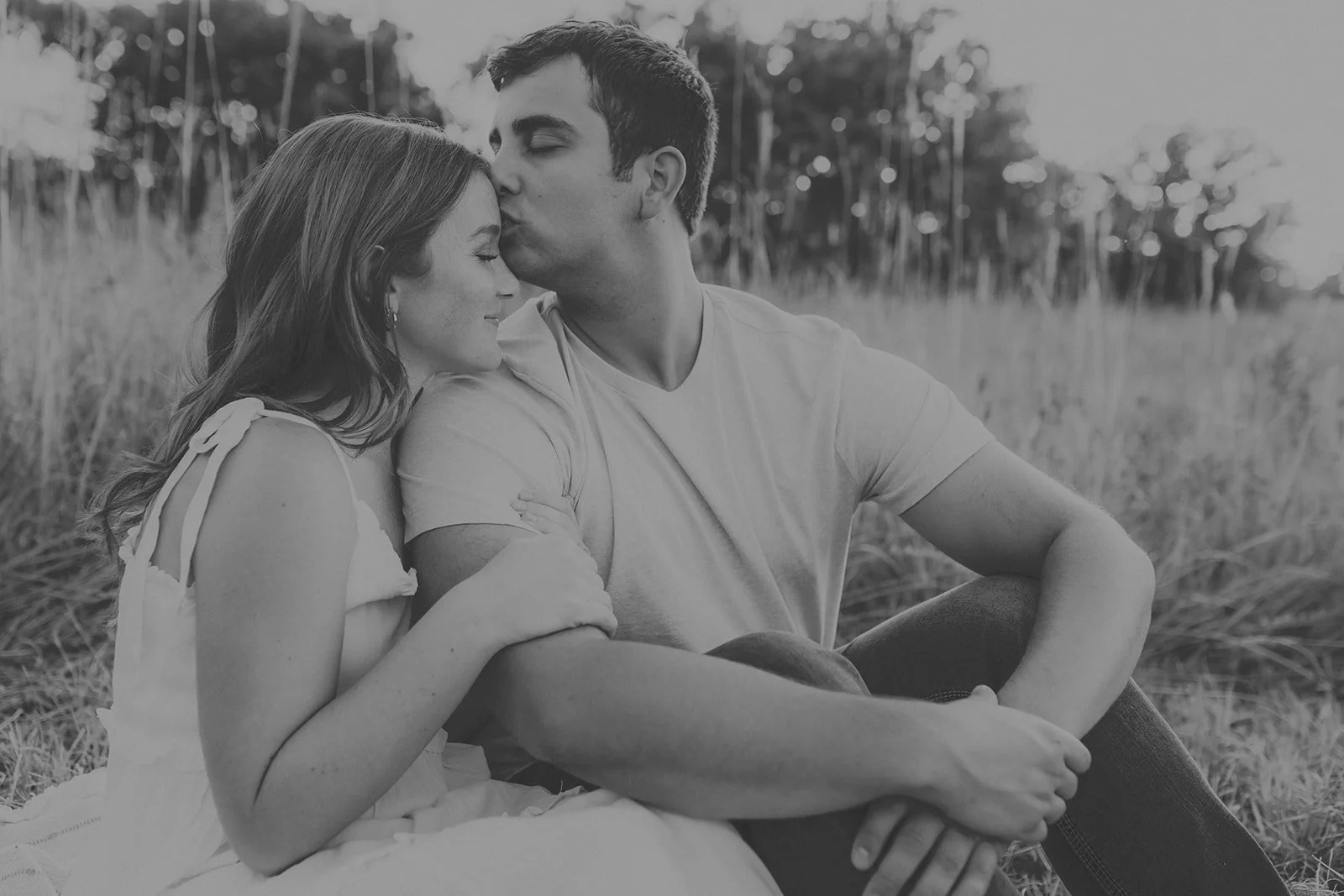 A black and white photo of a couple sitting outdoors in a field, with the man kissing the woman's forehead during their couples photography session at Rock Bridge State Park in Columbia, MO.