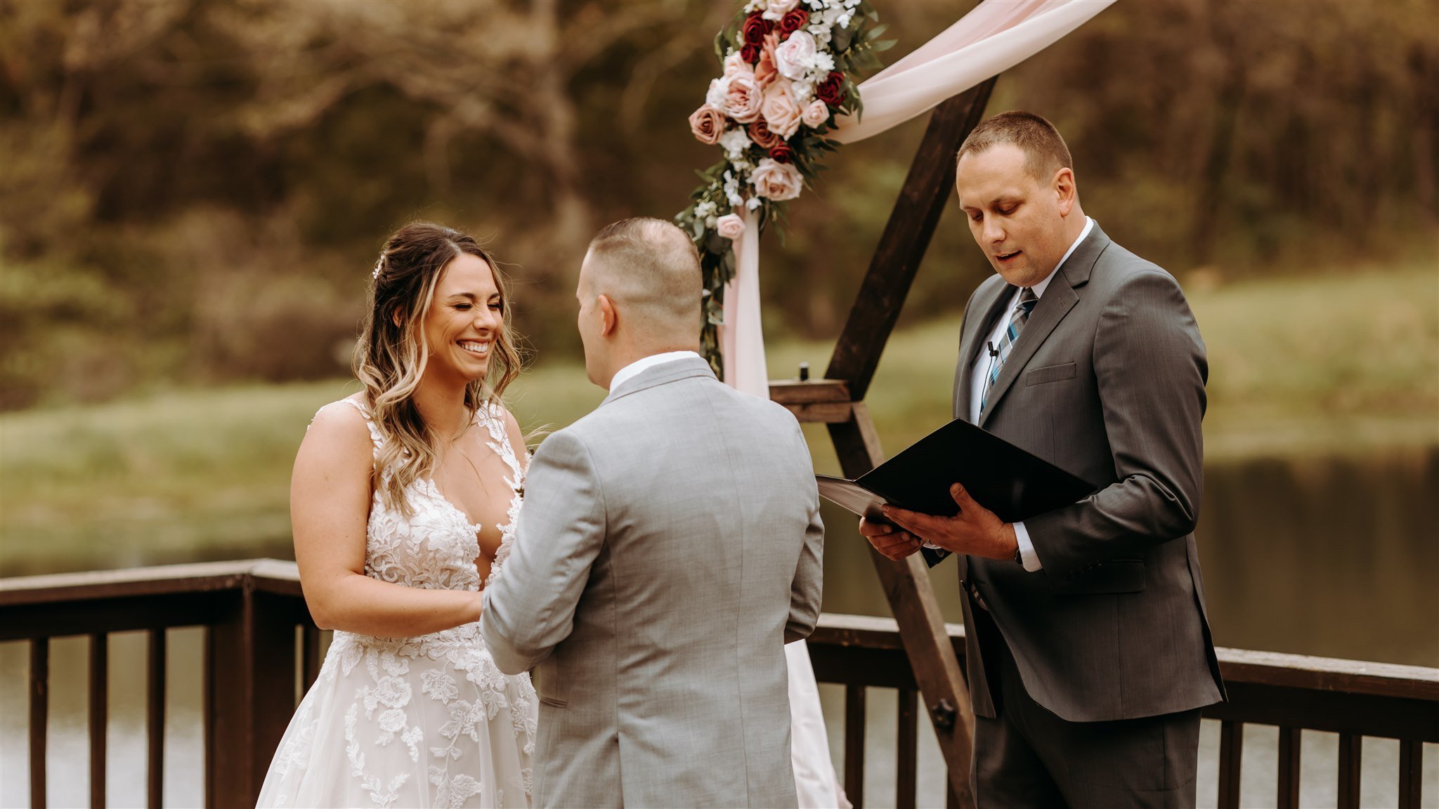 Bride and groom exchanging vows during outdoor wedding ceremony with officiant reading from a book, decorated with a floral arch, near a body of water and trees at Rolla, Missouri rustic wedding venue, The Village. Photos by Columbia, MO Photographer