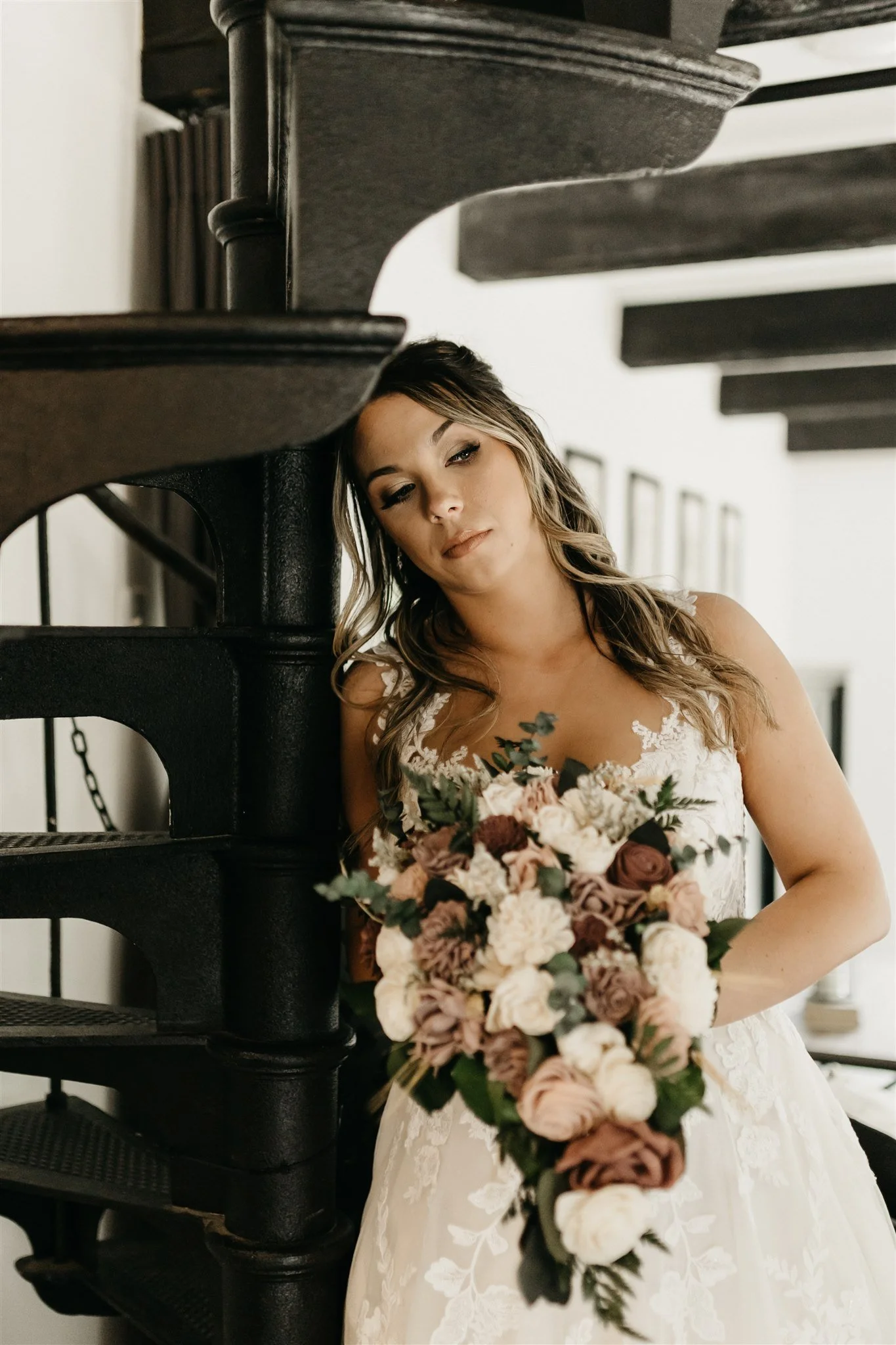 A bride in a white wedding dress holding a bouquet of roses and other flowers, standing next to a black spiral staircase at Rolla, Missouri rustic wedding venue, The Village. Photos by Columbia, MO Photographer Liv Strange Photography.