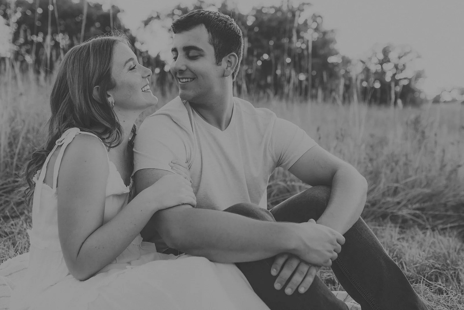 A black and white photo of a smiling young couple sitting in a field, looking at each other affectionately, with trees in the background during their couples photography session at Rock Bridge State Park in Columbia, MO.