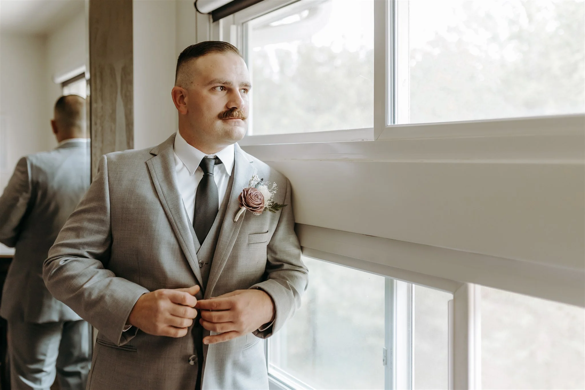 Portrait of groom dressed in a gray suit with a black tie and pink boutonniere, standing by a window looking outside, at Rolla, Missouri rustic wedding venue, The Village. Photos by Columbia, MO Photographer Liv Strange Photography.