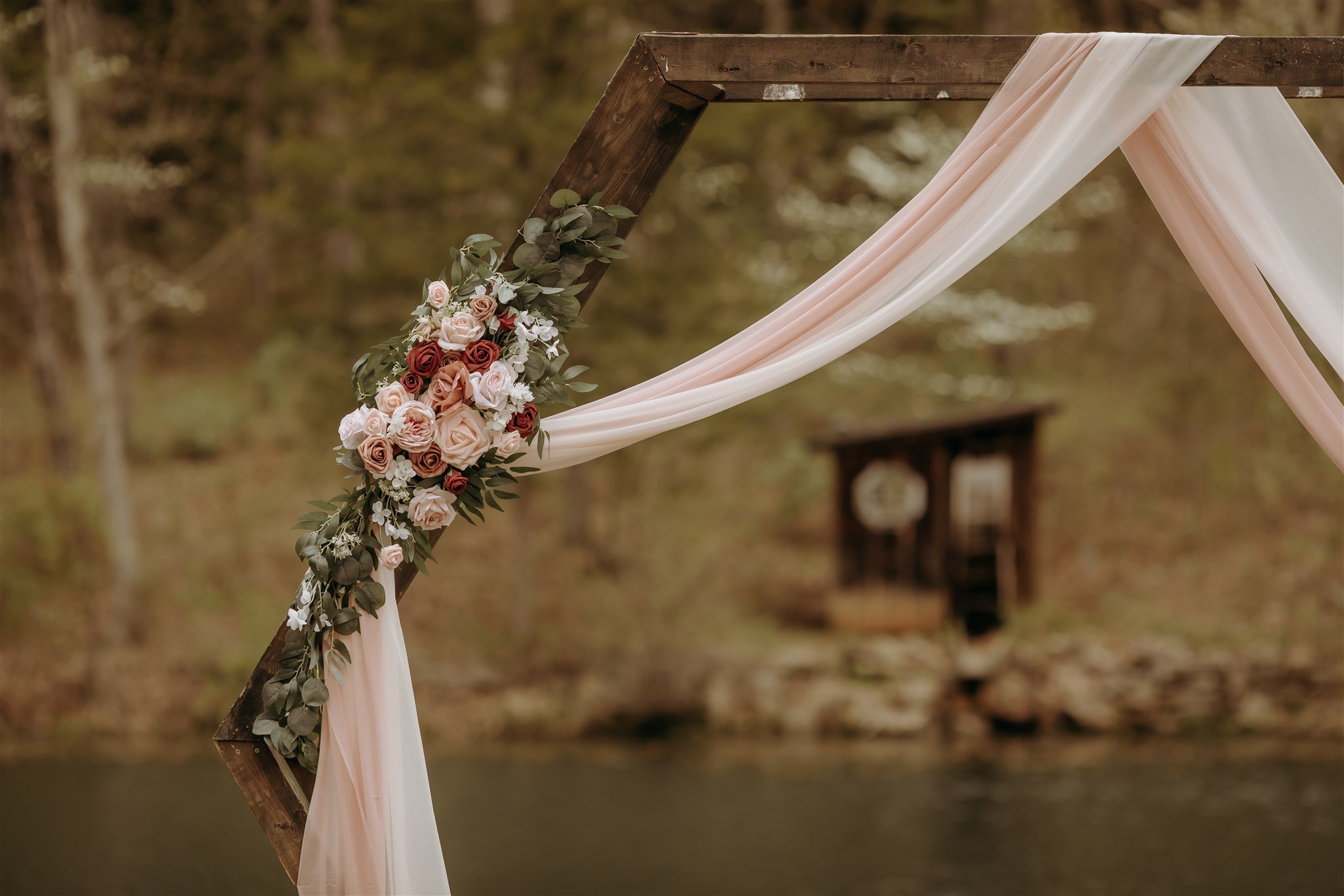 Wooden wedding arch with draped fabric and a floral arrangement of roses and greenery, outdoors near a lake with trees in the background at Rolla, MO rustic wedding venue, The Village.