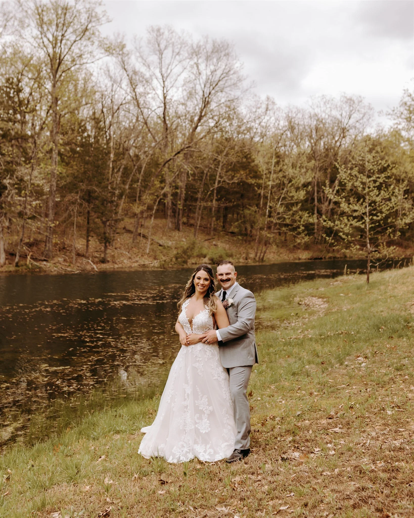 A bride and groom standing together by a river, surrounded by trees with fall foliage at Rolla, MO rustic wedding venue, The Village. Photos by Columbia, MO Photographer Liv Strange Photography.
