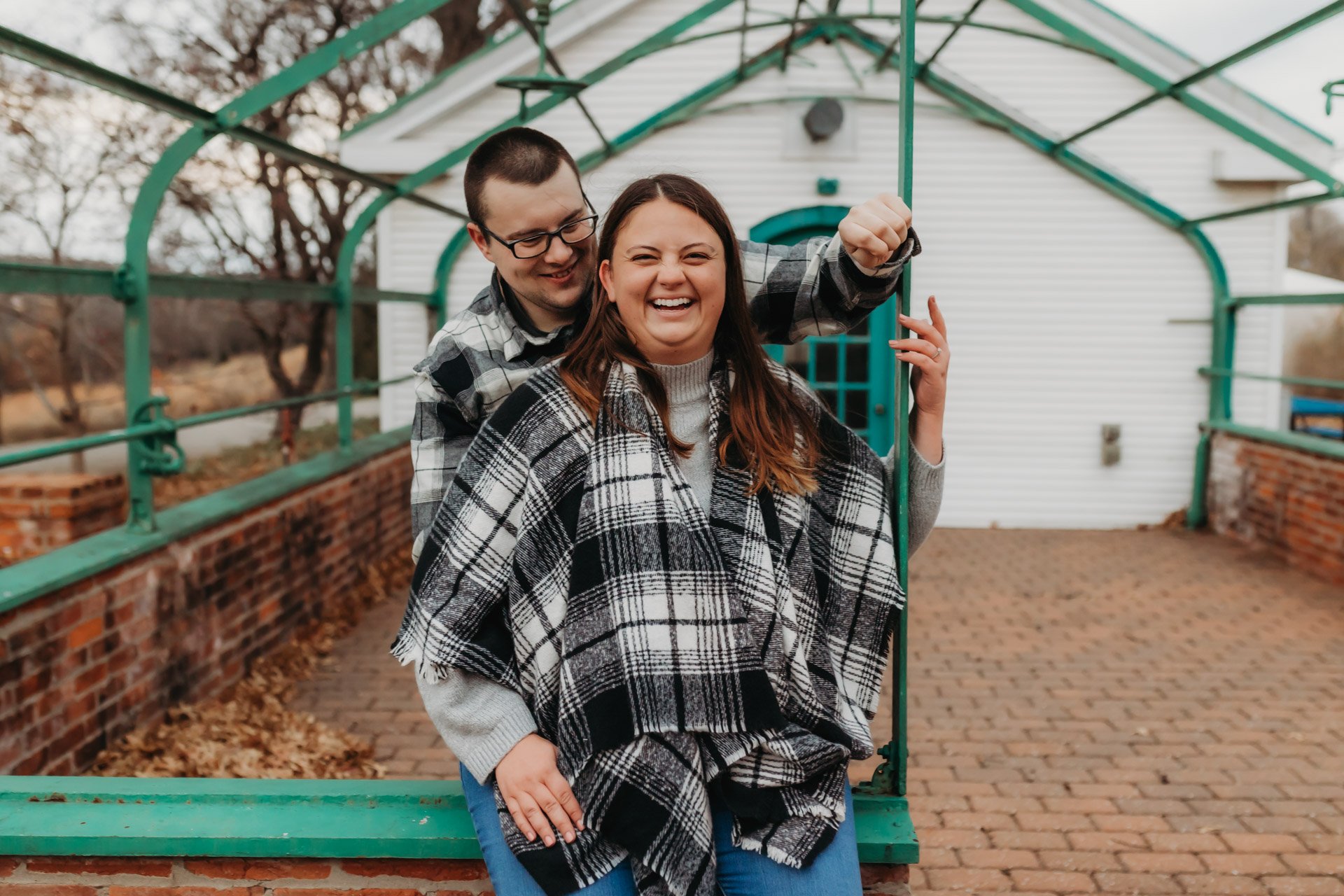 A smiling couple standing outdoors on a brick patio with a small white house in the background, sharing a playful moment under a green metal structure at their engagement photography session at Queeny Park in STL.