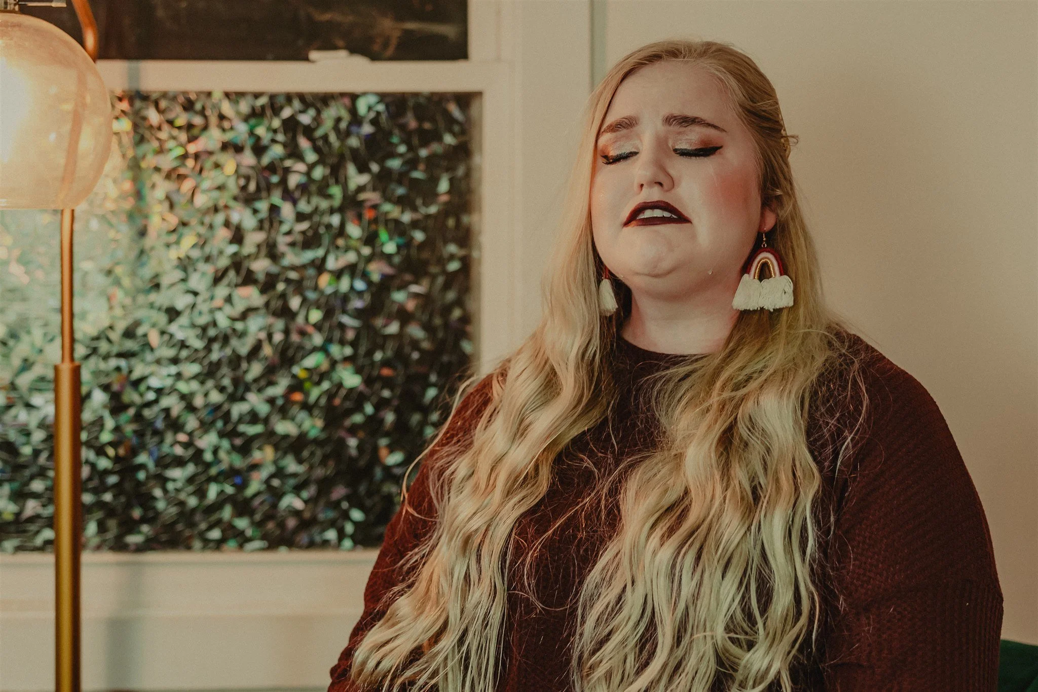 A woman with long wavy blonde hair and makeup, wearing rainbow-shaped tassel earrings, dark lipstick, and a maroon sweater, appears emotional with eyes closed and tears on her face during a trauma informed photography session in Columbia, Missouri.