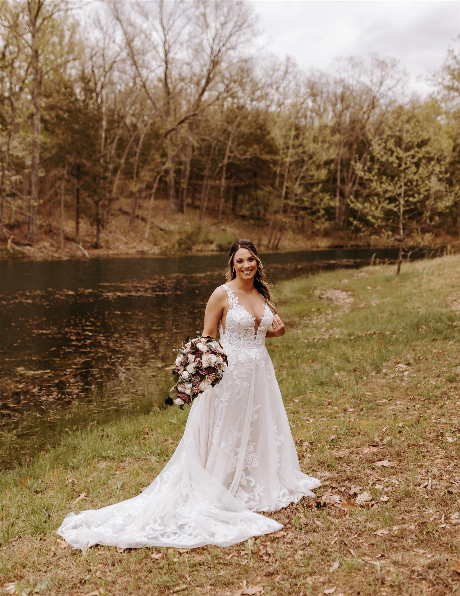 A bride in a white wedding dress holding a bouquet, standing on grass near a river surrounded by trees at Rolla, MO rustic wedding venue, The Village. Photos by Columbia, MO Photographer Liv Strange Photography.