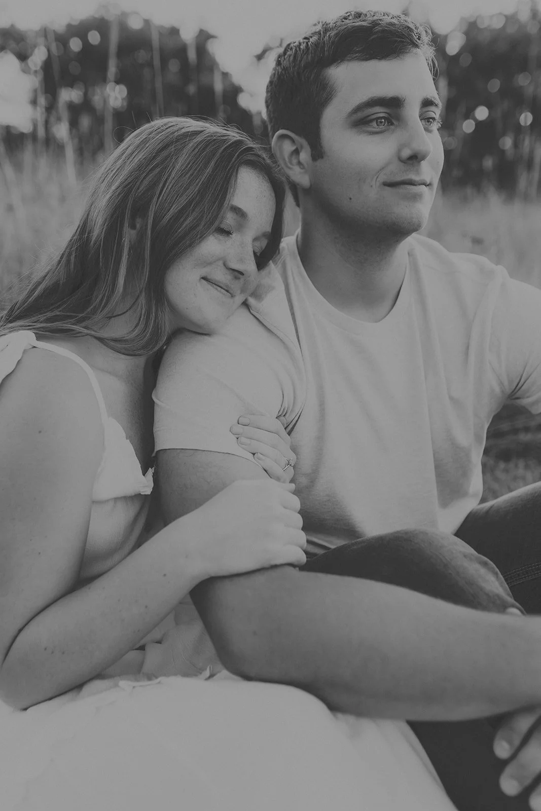 A smiling woman leaning her head on a man’s shoulder, both sitting outdoors, with trees in the background, in black and white during their engagement photography session at Rock Bridge State Park in Columbia, MO.