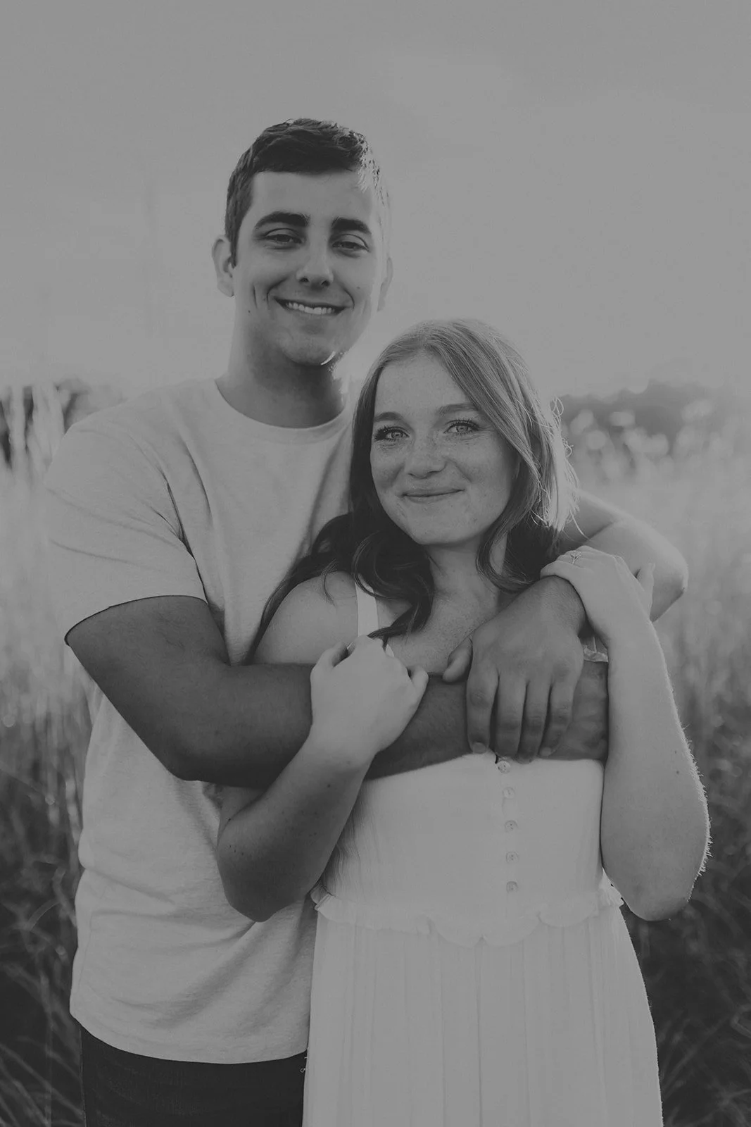 A smiling young man standing behind a young woman, both with arms around each other, outdoors in a field with tall grass, in black and white during their engagement photography session at Rock Bridge State Park in Columbia, MO.