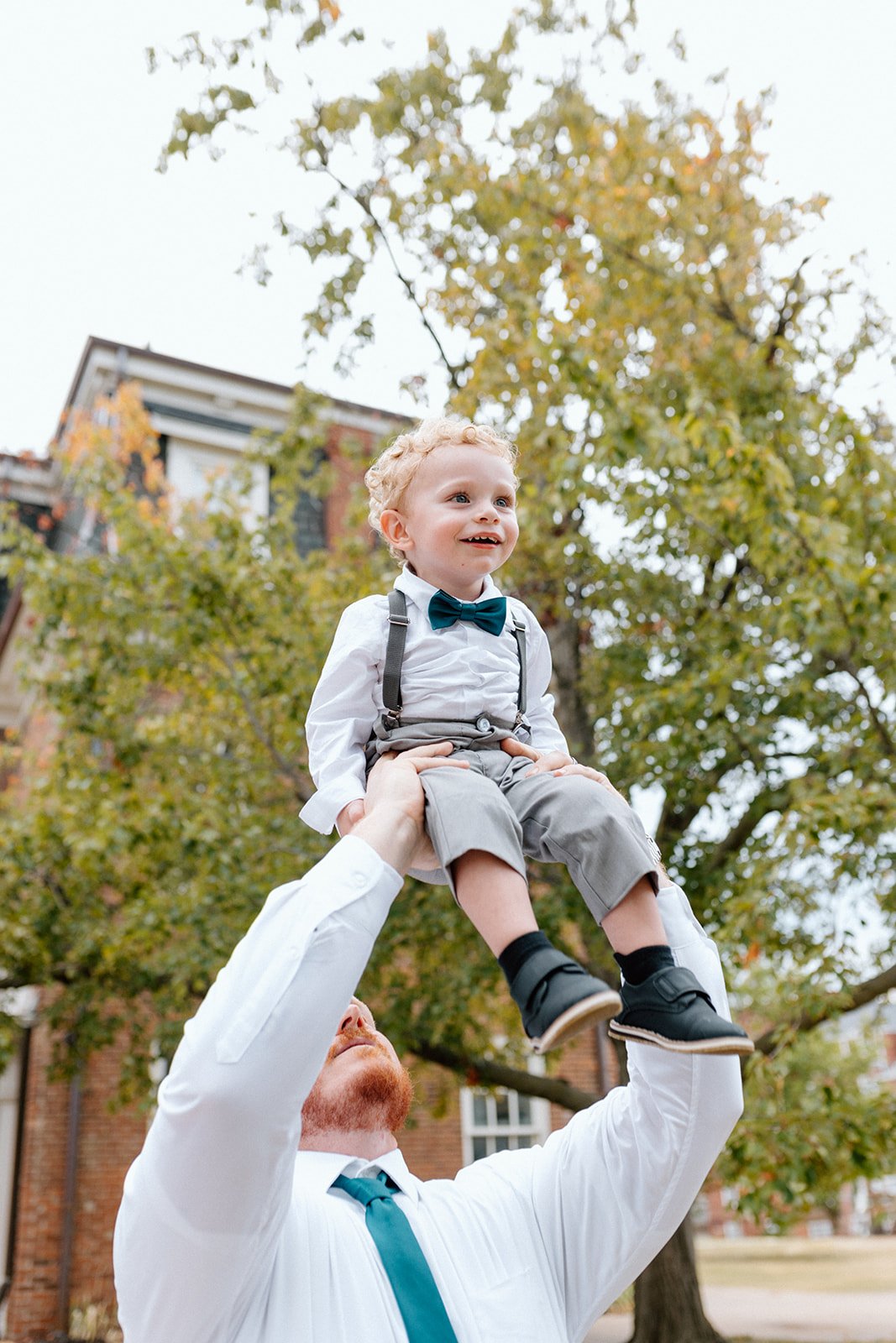 A man with red hair and a white shirt lifts a young boy with curly blonde hair into the air outdoors. The boy, dressed in a white shirt, gray pants, suspenders, and a blue bow tie, smiles happily at an LGBTQ+ wedding in Columbia, MO at wedding venue,