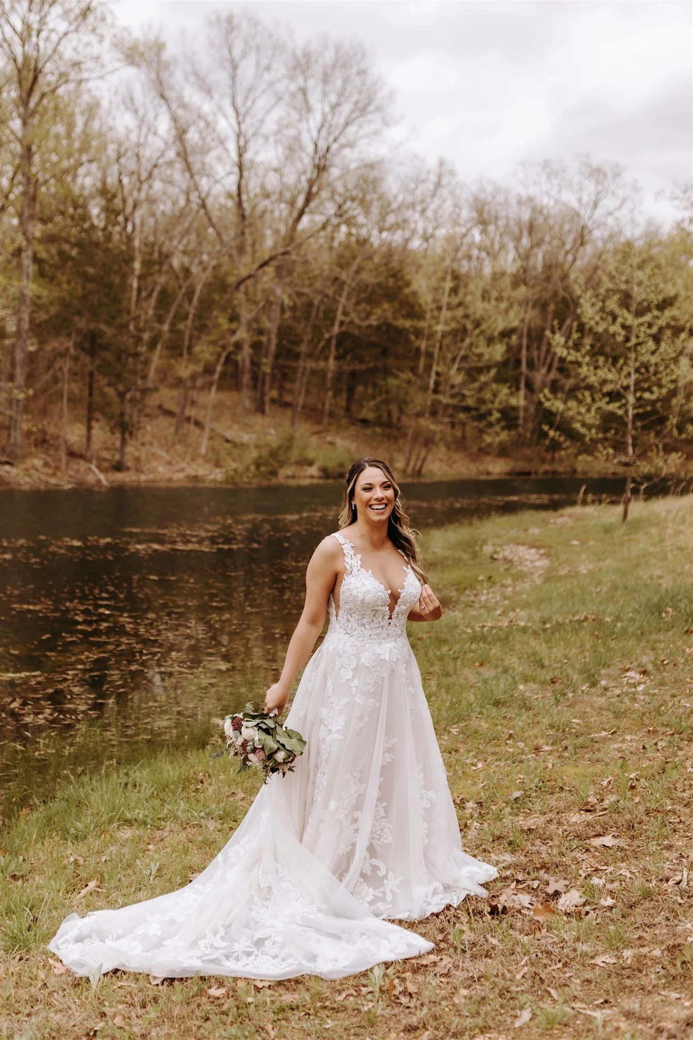 A bride in a white lace wedding dress, holding a bouquet, standing beside a pond with trees in the background at Rolla, MO rustic wedding venue, The Village. Photos by Columbia, MO Photographer Liv Strange Photography.