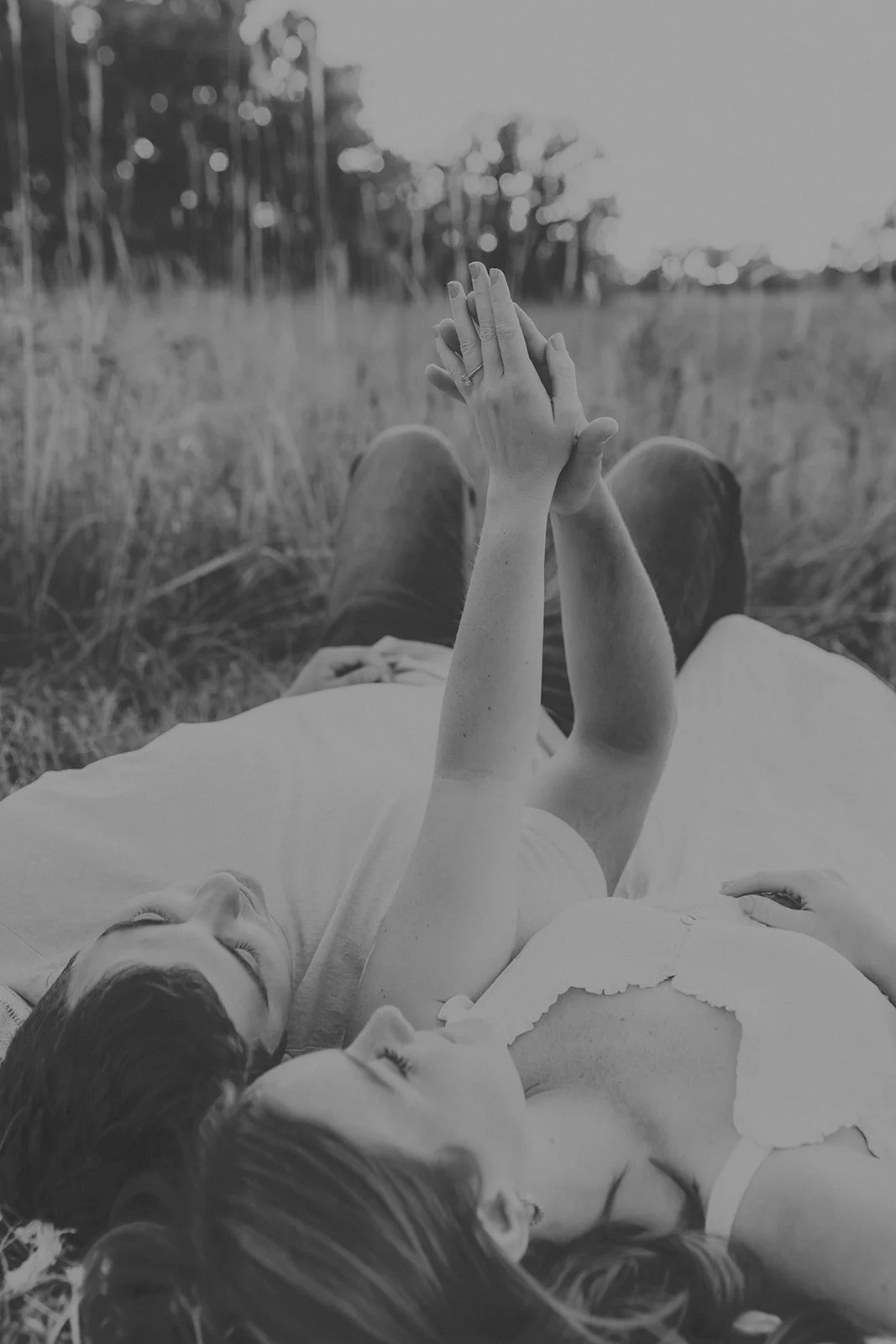A young engaged couple lying on grass in a field, holding hands and touching fingers, with trees in the background, black and white photo at couples photography session at Rock Bridge State Park in Columbia, MO.